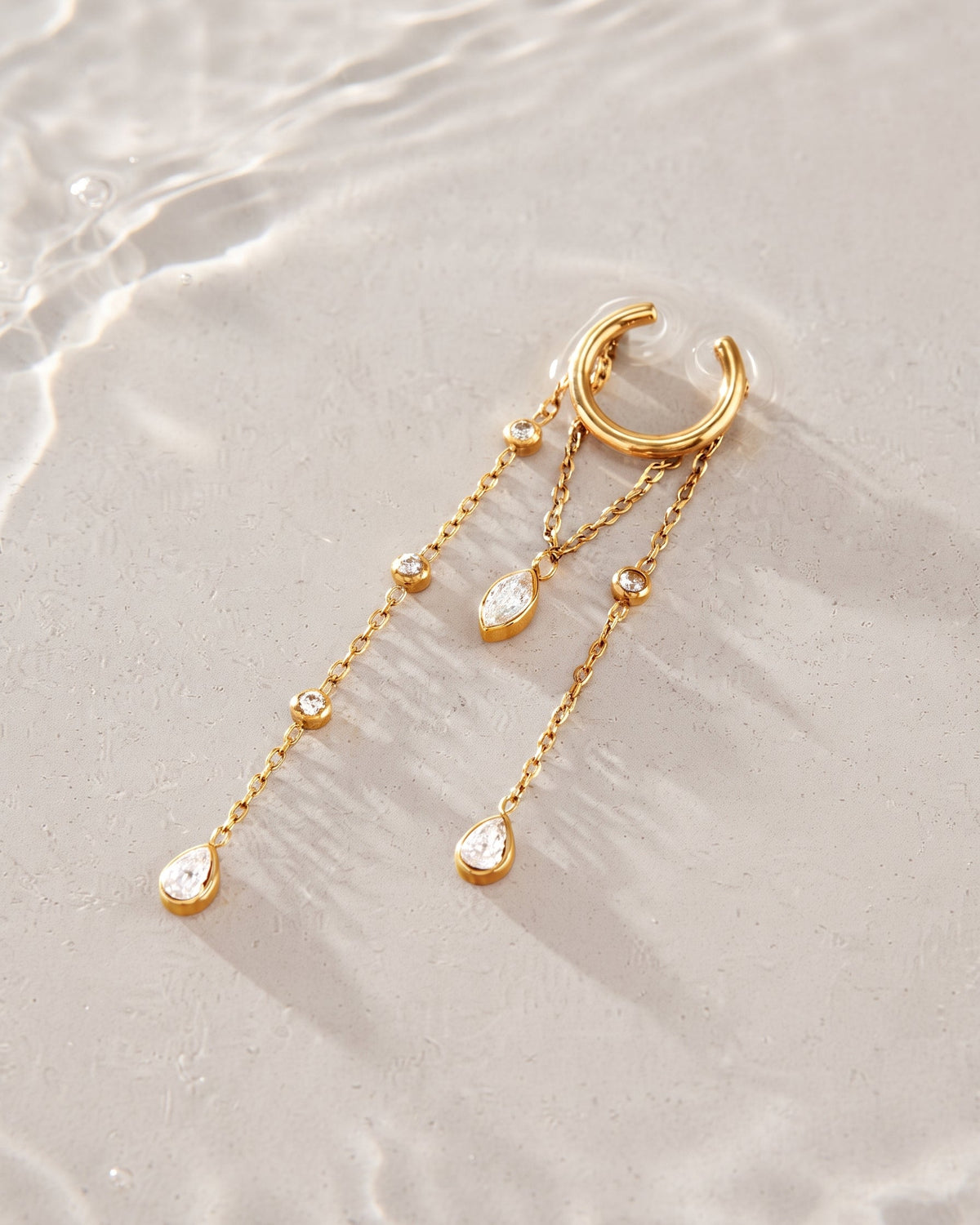 A gold crescent-shaped hoop earring with three dangling chain drops and teardrop crystal charms, styled on a light beige sandy surface with soft water ripples in the background.