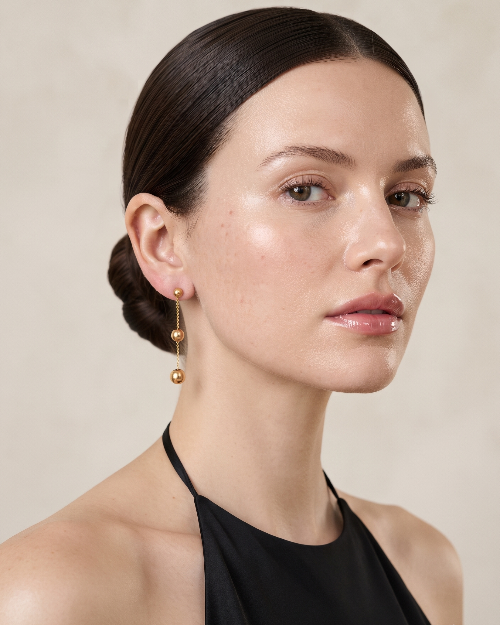 A woman with her hair in a low bun wears a pair of gold drop earrings with spherical beads, photographed against a soft beige background.