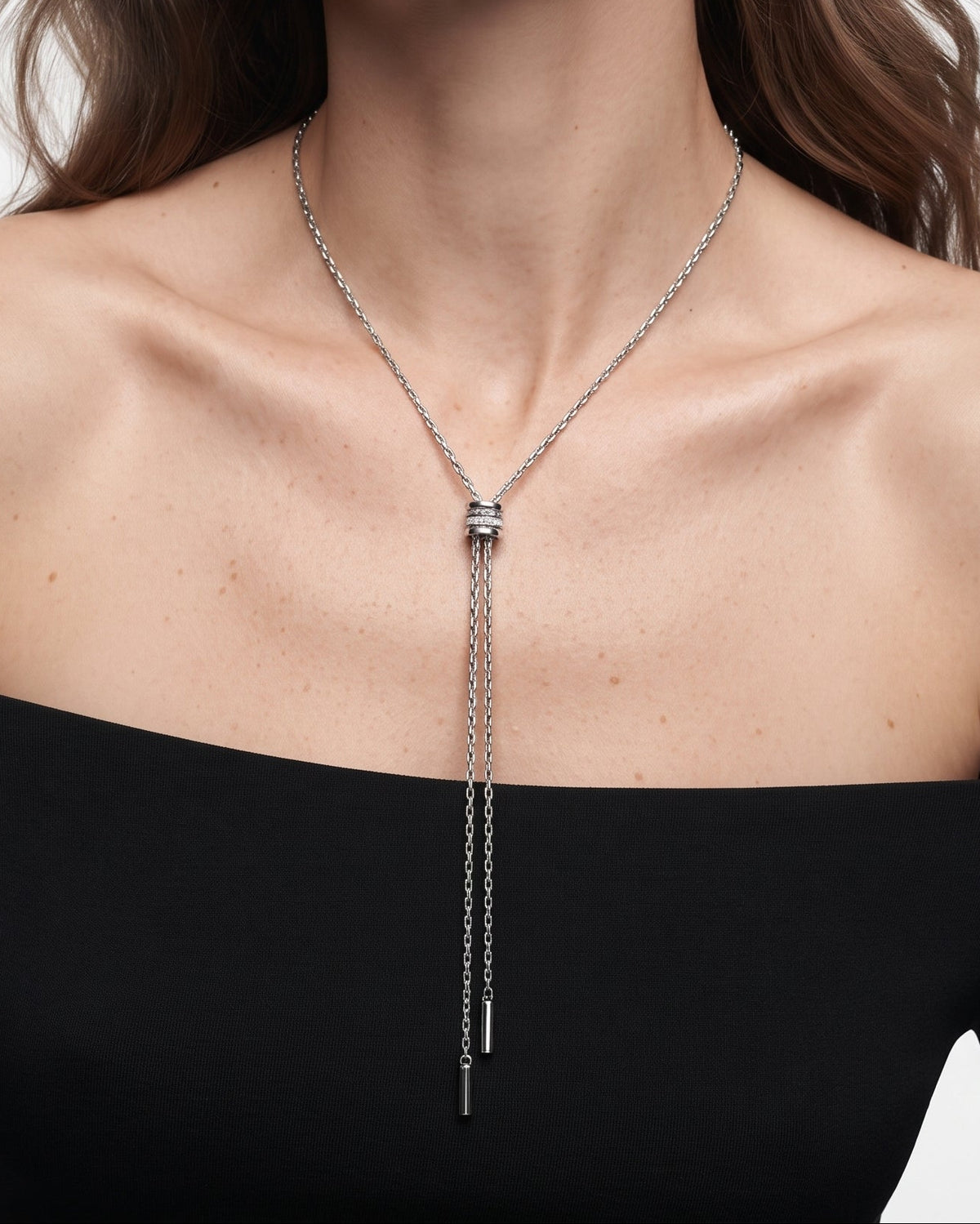 A woman wearing a delicate silver lariat-style necklace with a central cylindrical connector and two long dangling chains, paired with a black off-the-shoulder top against a light gray studio background.