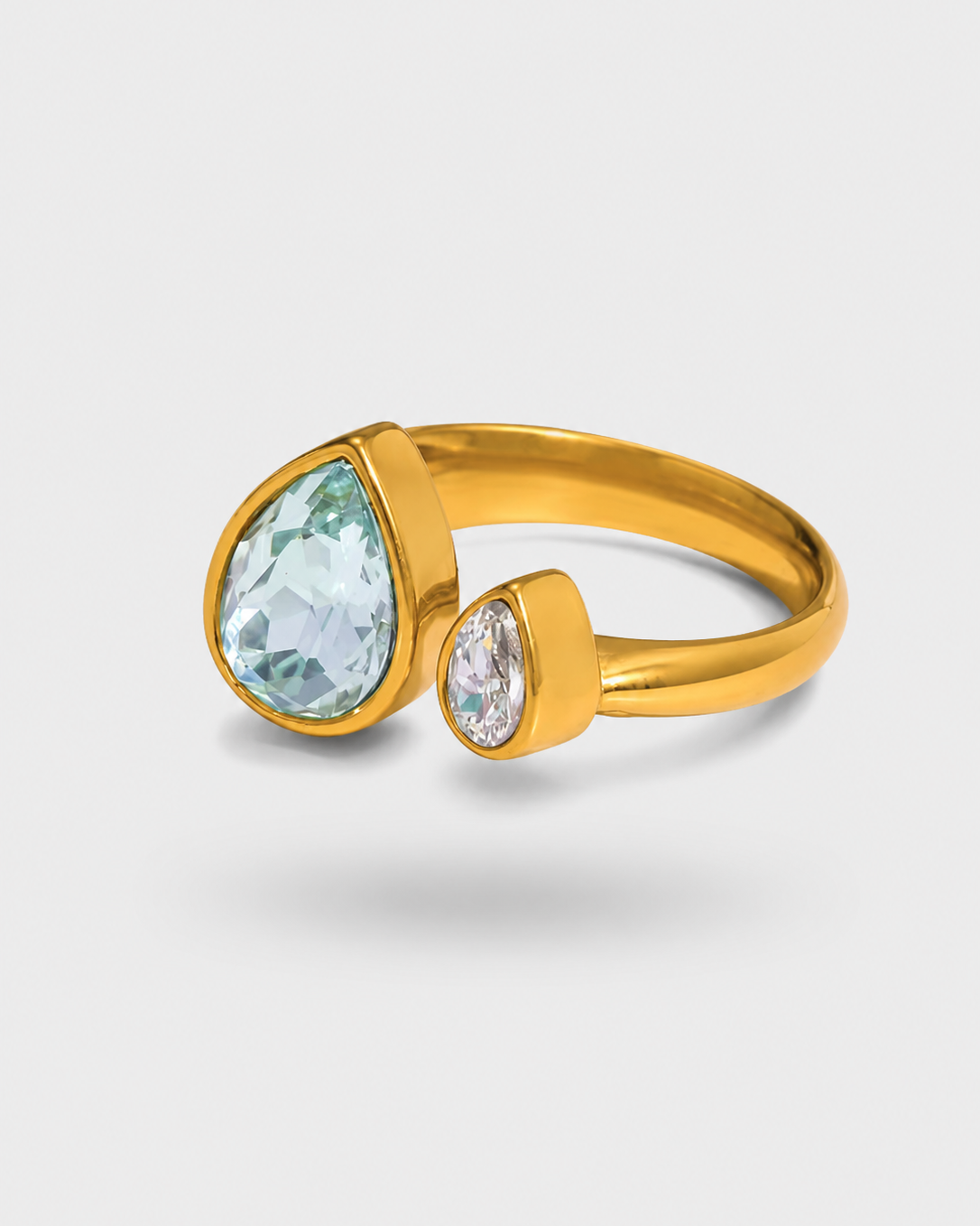 A gold ring featuring a large teardrop-shaped light blue gemstone and a smaller teardrop-shaped clear gemstone, set on a light gray background.