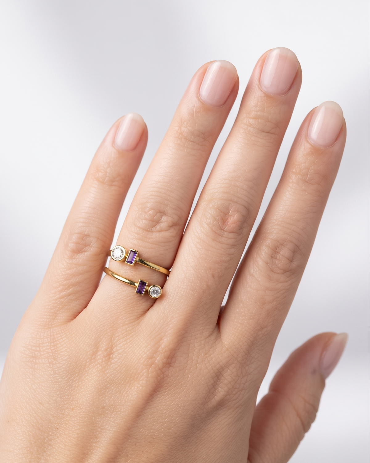 A close-up of a hand wearing two delicate gold rings with small clear gemstones and purple accents, set against a soft light gray background.