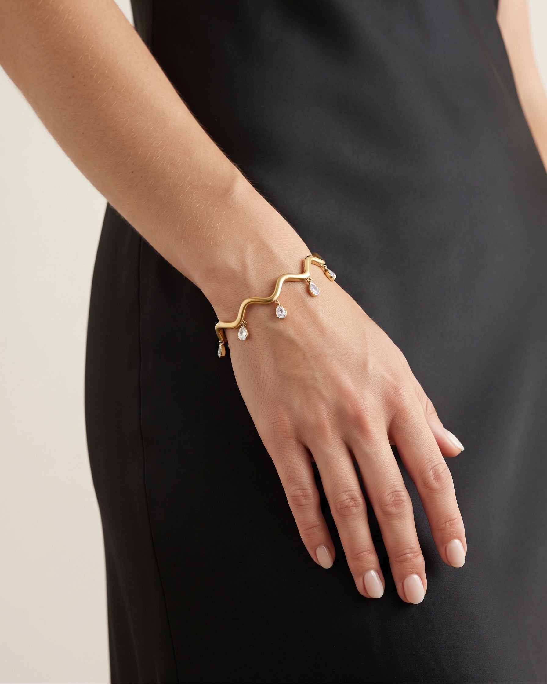 A gold wavy cuff bracelet with small clear teardrop gemstones, worn on a model's wrist. The model is wearing a black satin dress against a light beige background.