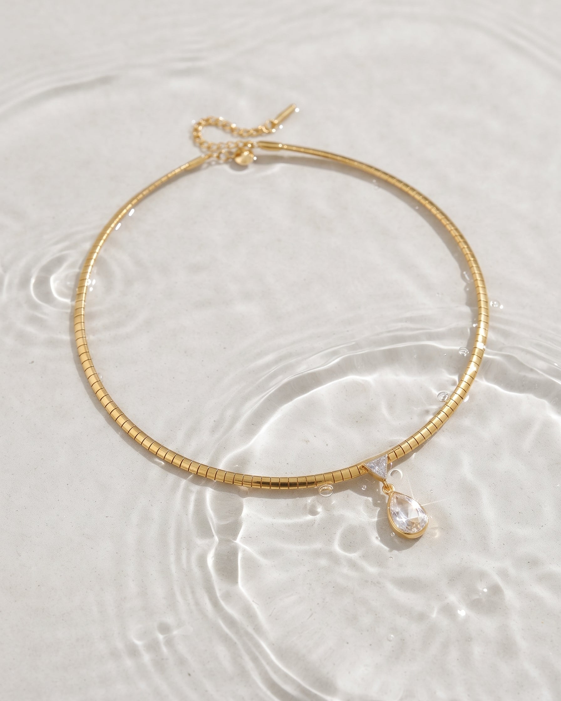 A gold-toned snake chain necklace with a small clear teardrop crystal pendant, resting on clear water with ripples.