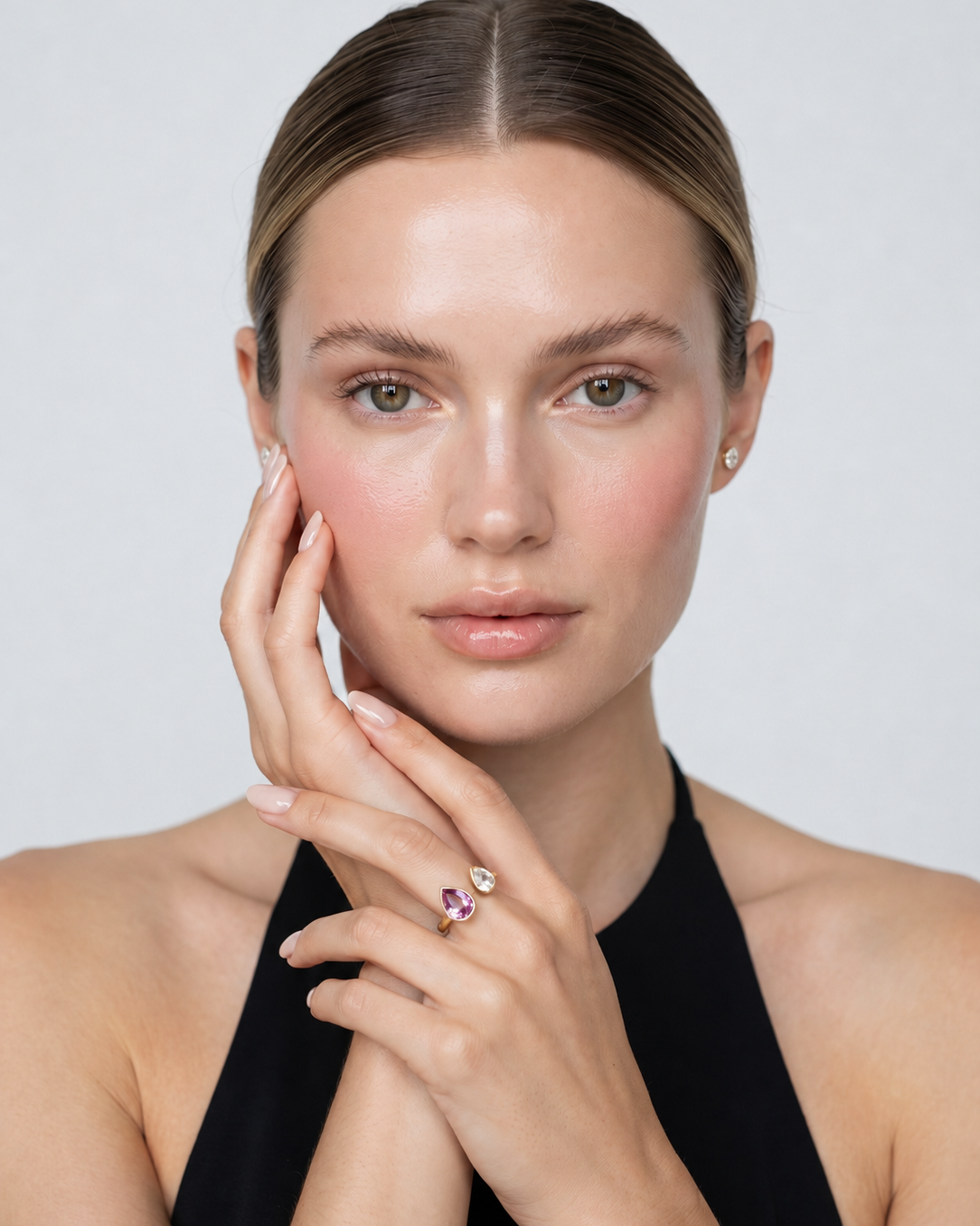 A close-up portrait of a woman wearing a delicate ring with a small purple heart and a silver heart on a thin band, set against a clean light gray background.