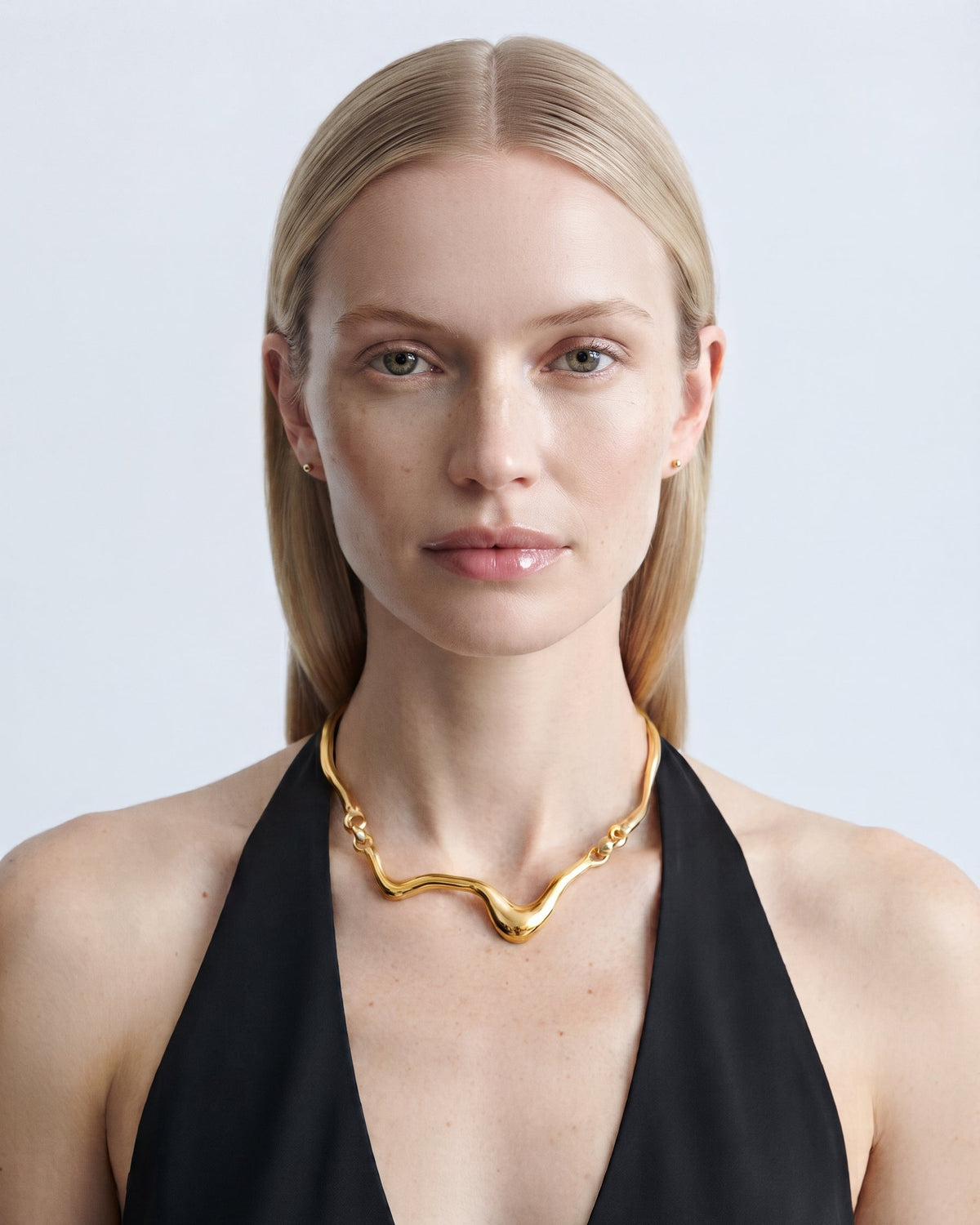 A blonde model wearing a wavy gold chain necklace and a black halter top, photographed against a light gray studio background.