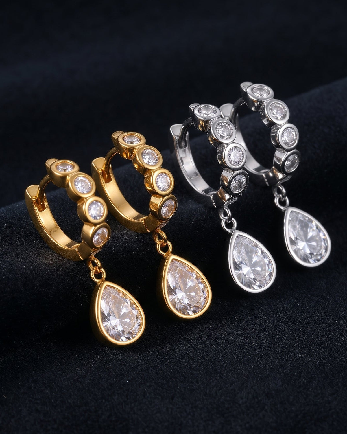 A pair of gold huggie hoop earrings with clear teardrop gemstones, displayed alongside a matching pair of silver huggie hoop earrings on a dark textured fabric background.
