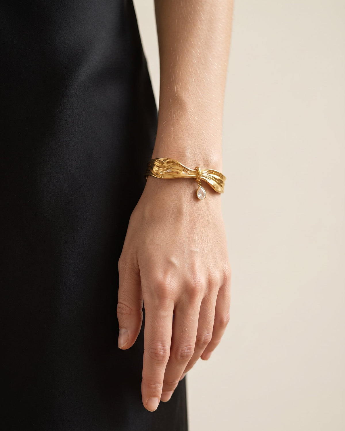 A close-up of a person's hand and wrist wearing a textured gold bow-shaped cuff bracelet with a small clear teardrop gemstone, set against a dark black dress and a light beige background.