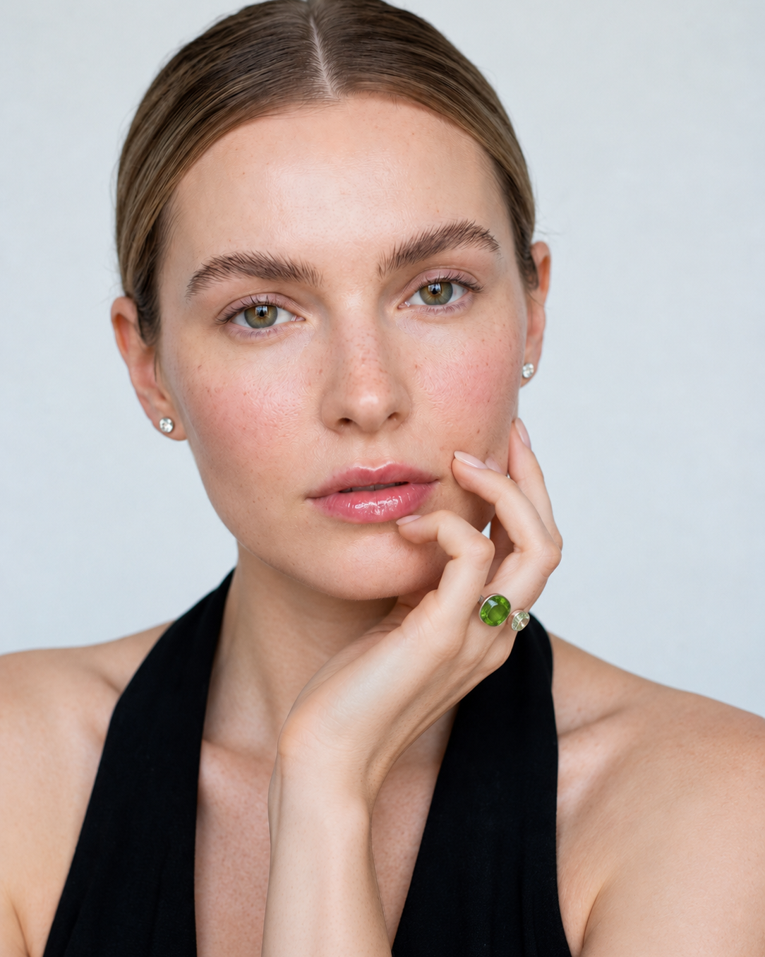 A close-up portrait of a woman with her hair pulled back, wearing a black halter top and a green gemstone ring on her finger, against a light gray background.