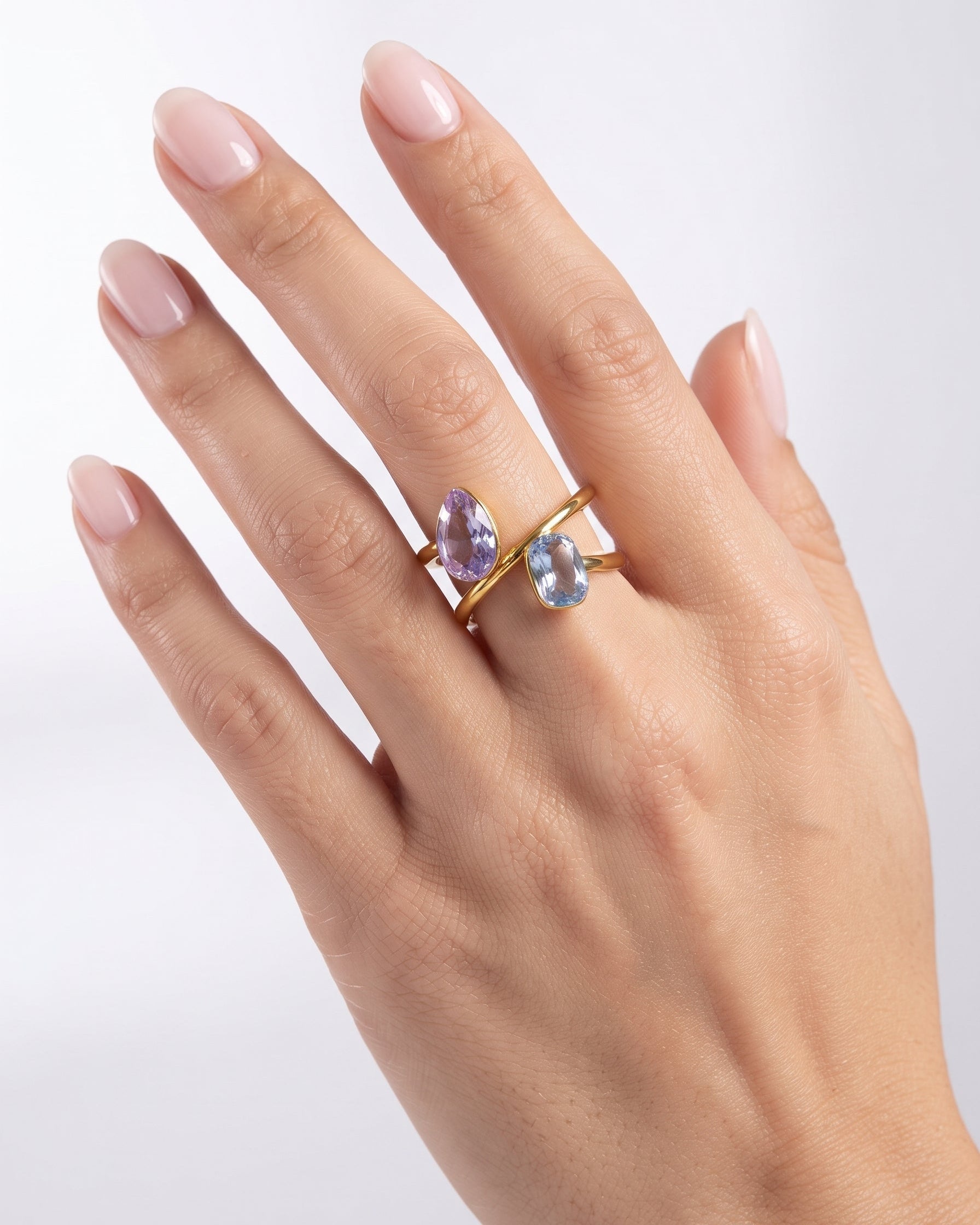 A close-up of a hand with pale pink manicured nails wearing a gold ring with two teardrop-shaped gemstones, one light purple and one light blue, set against a clean white background.