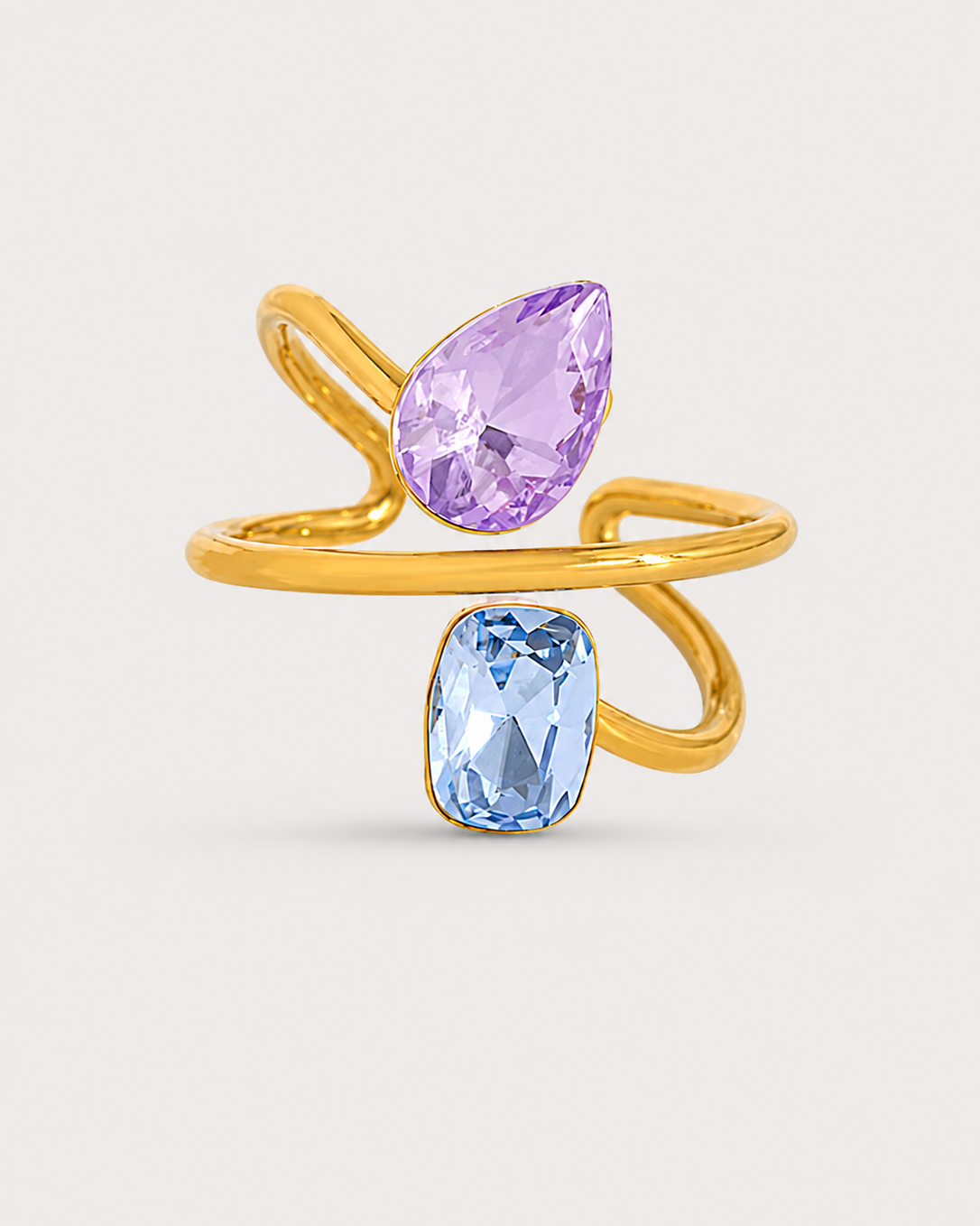 A gold wrap ring featuring a large teardrop-shaped light purple gemstone on the top curve and a smaller rectangular light blue gemstone on the bottom curve, set against a light beige background.