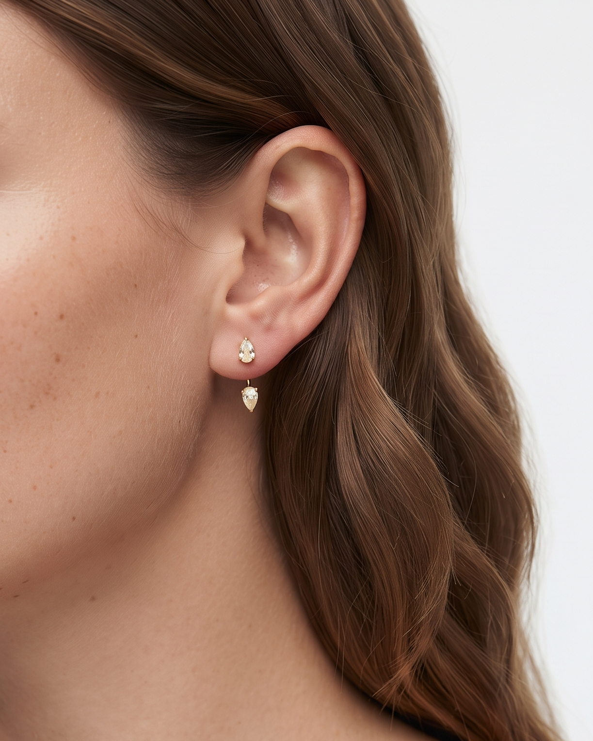 A close-up side profile of a woman wearing a delicate gold stud earring with a small teardrop-shaped gold charm, set against a clean white background.