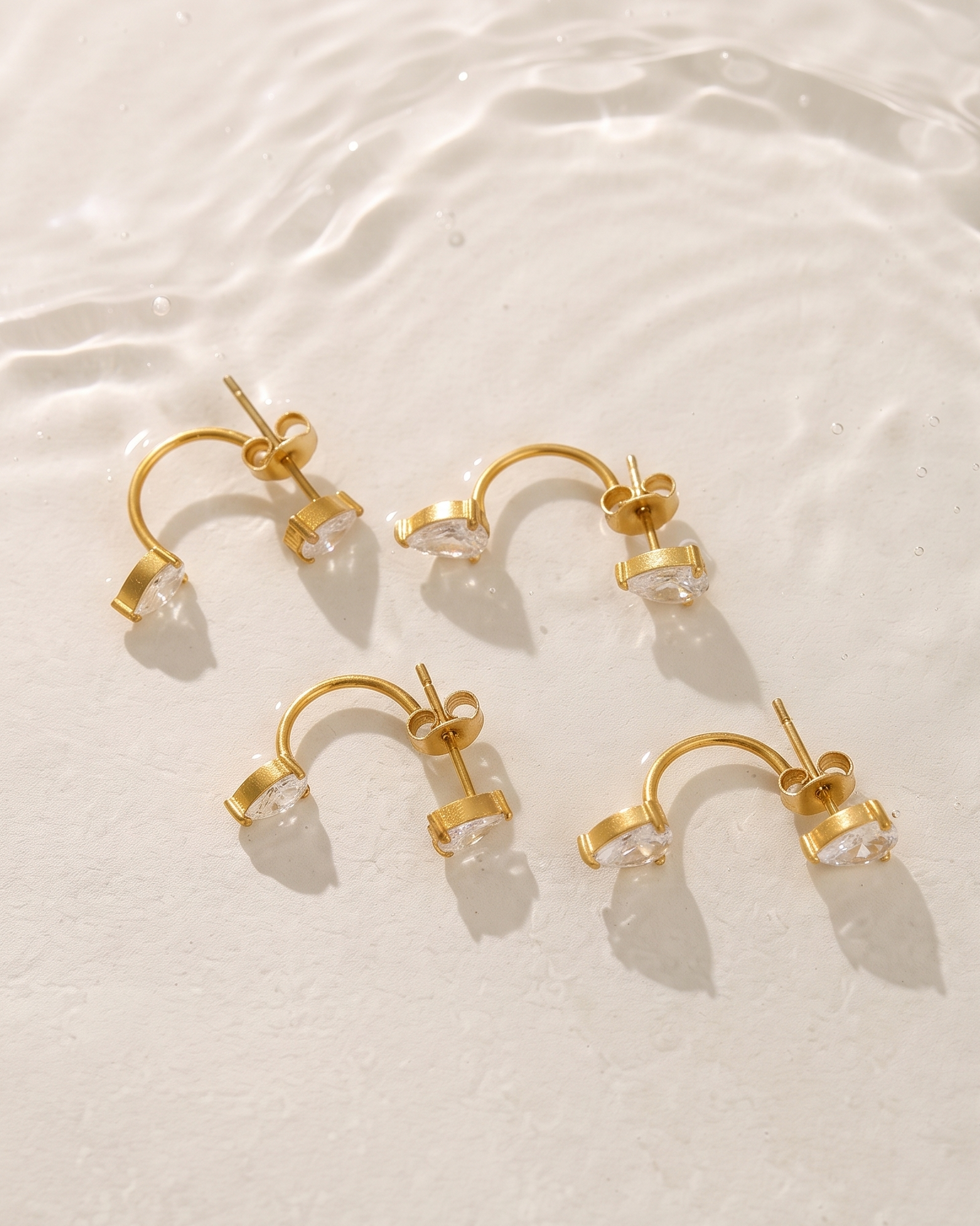Four gold crescent-shaped hoop earrings with clear faceted gemstones, arranged on a textured white surface with soft water ripples.