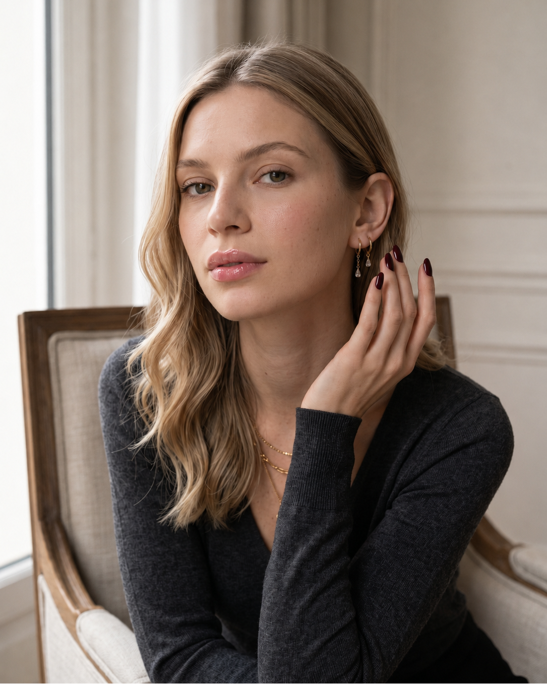 A woman with blonde wavy hair sits in a light-colored armchair, wearing a dark charcoal gray long-sleeve top and delicate gold jewelry, including a necklace and earrings.