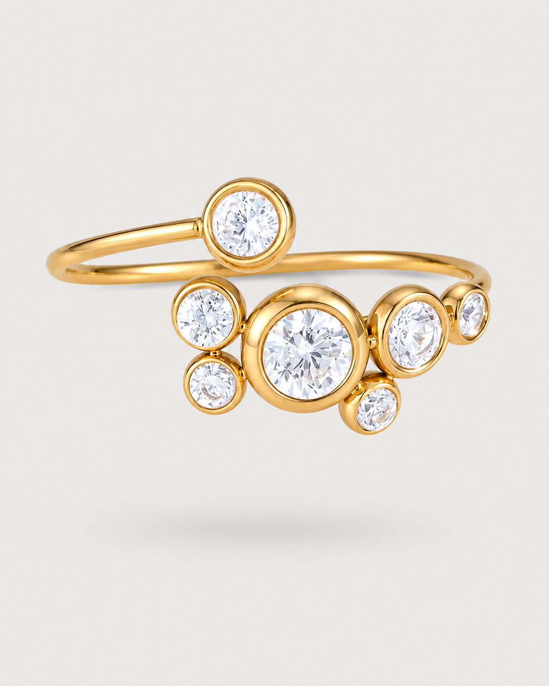 A gold ring featuring a curved band with multiple bezel-set round clear gemstones in varying sizes, displayed on a light gray background.