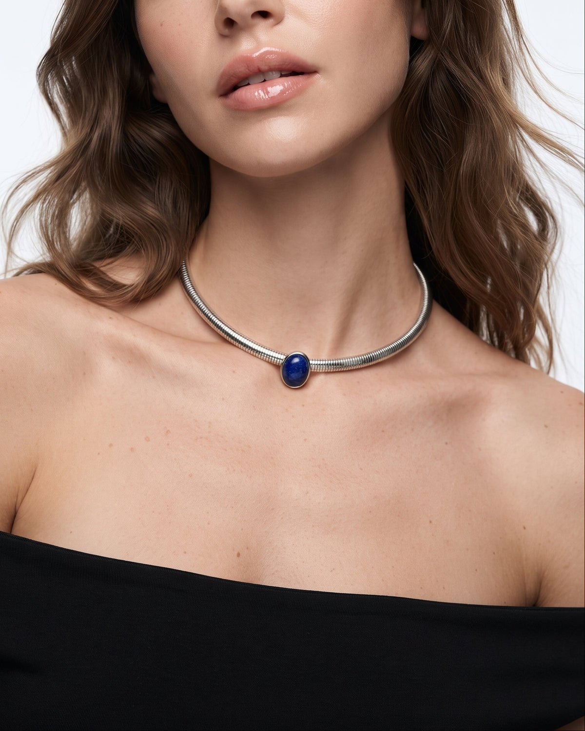 A close-up of a woman wearing a silver-toned metal choker necklace with a central deep blue oval stone, paired with a black off-the-shoulder top against a light gray background.
