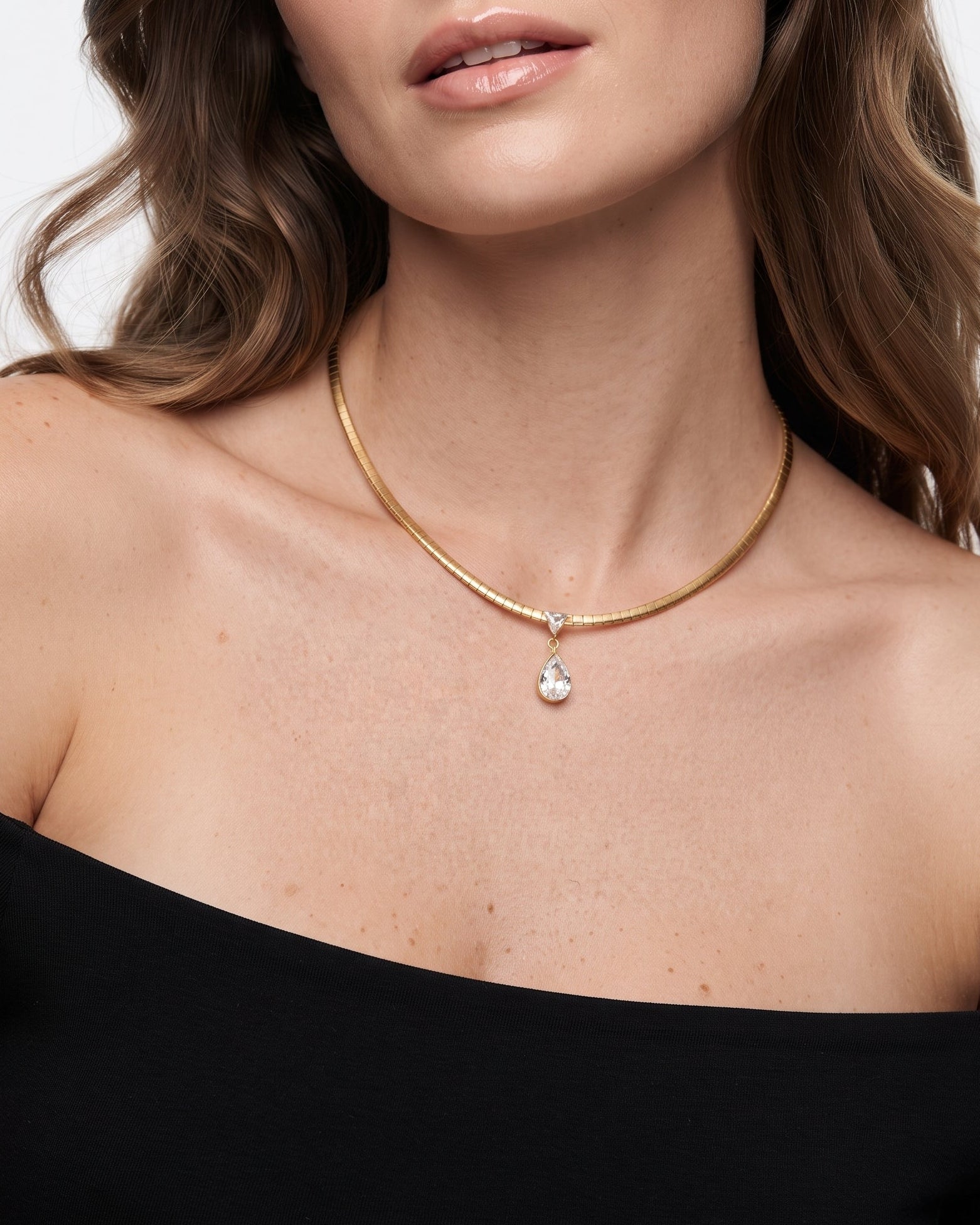 A close-up of a woman wearing a delicate gold chain necklace with a small teardrop-shaped clear gemstone pendant, styled with a black off-the-shoulder top against a light gray background.