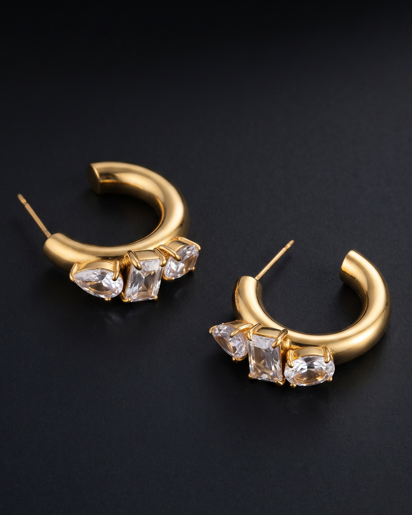 A pair of gold crescent-shaped hoop earrings with three clear rectangular gemstones on each earring, displayed on a dark textured background.