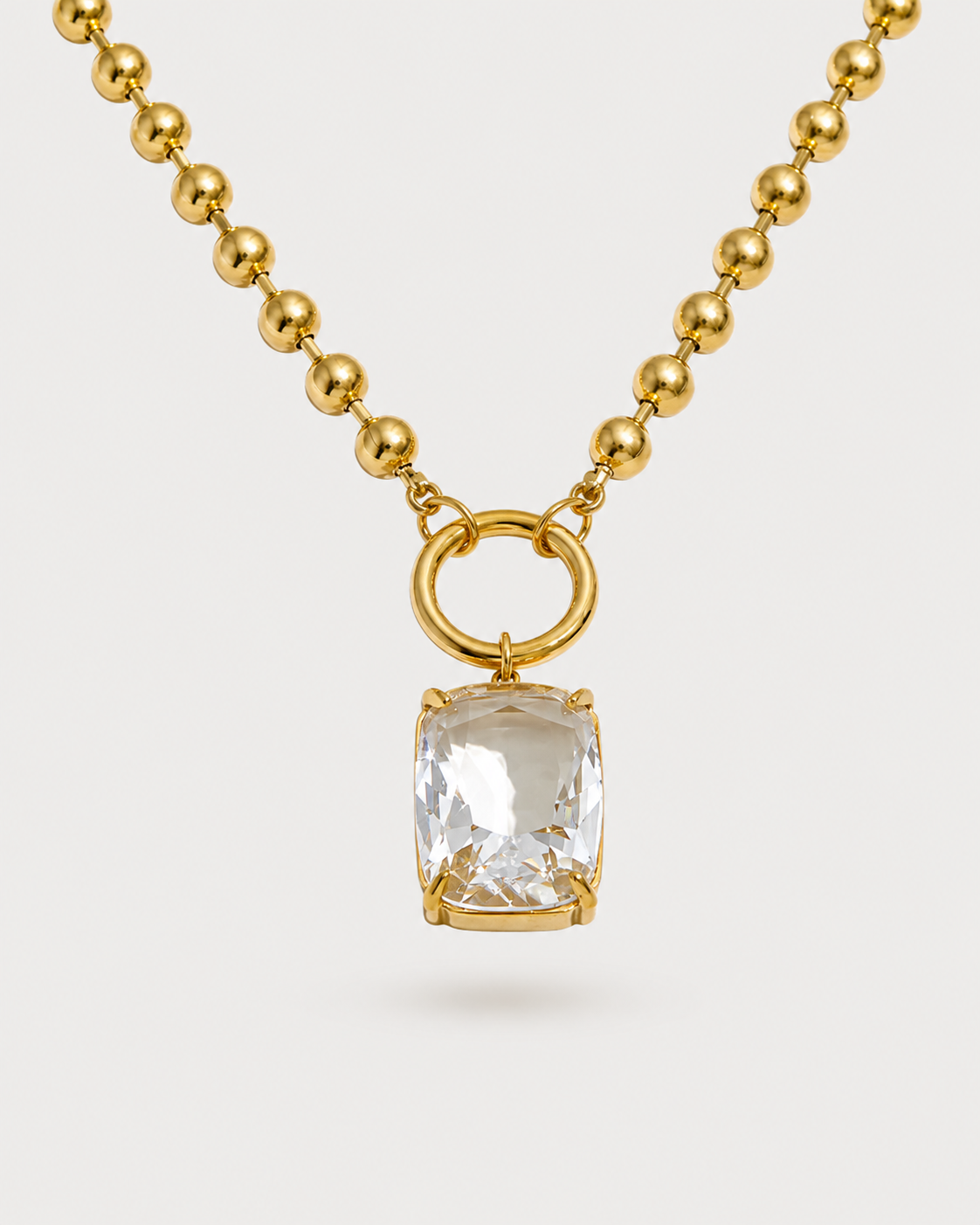 A gold ball-chain necklace featuring a cushion-cut clear gemstone pendant in a gold bezel setting, suspended from a gold ring connector, shown against a light gray background.