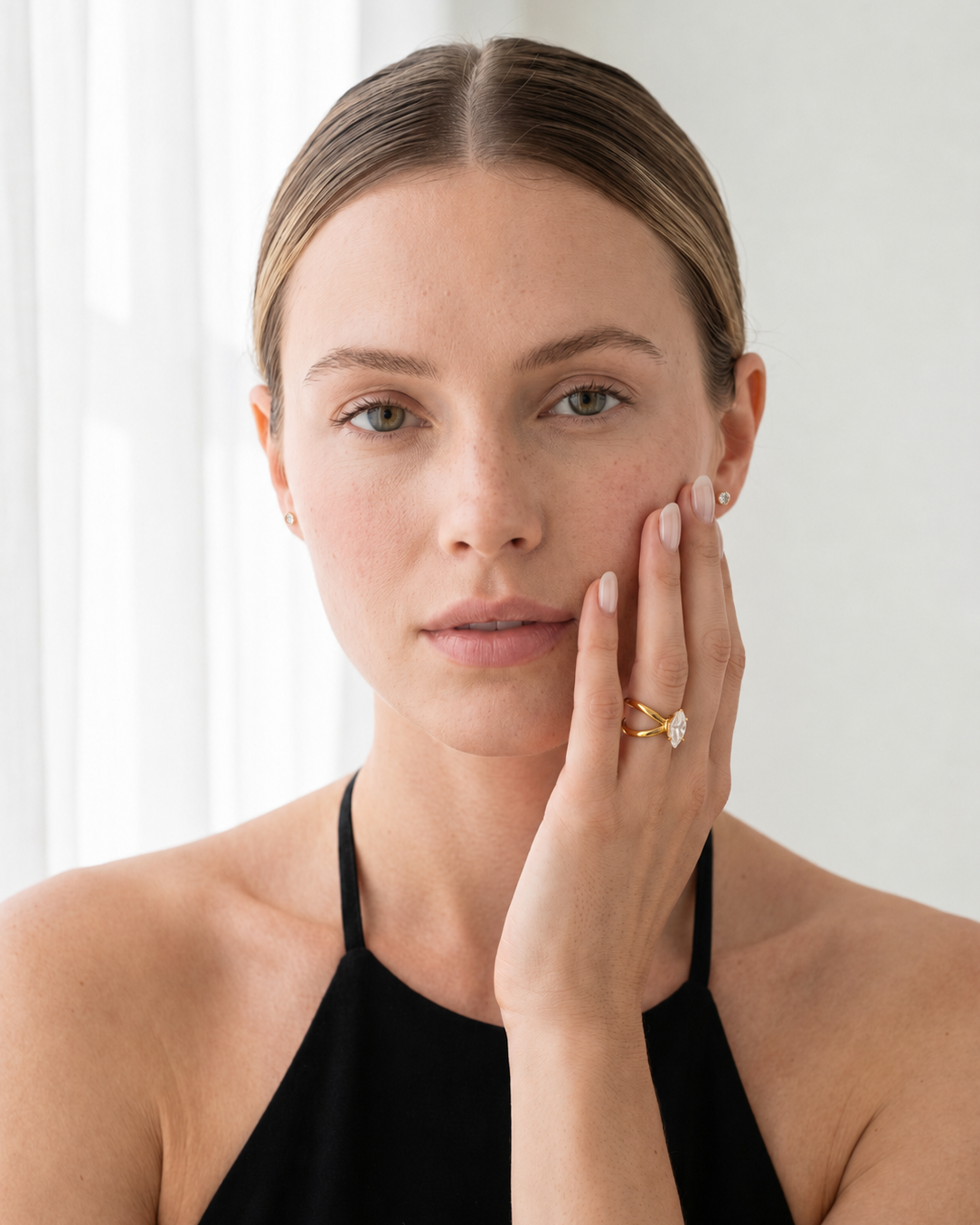 A woman with her hair pulled back wears a delicate gold ring with a small clear gemstone on her finger, posing with her hand against a bright, softly lit background.