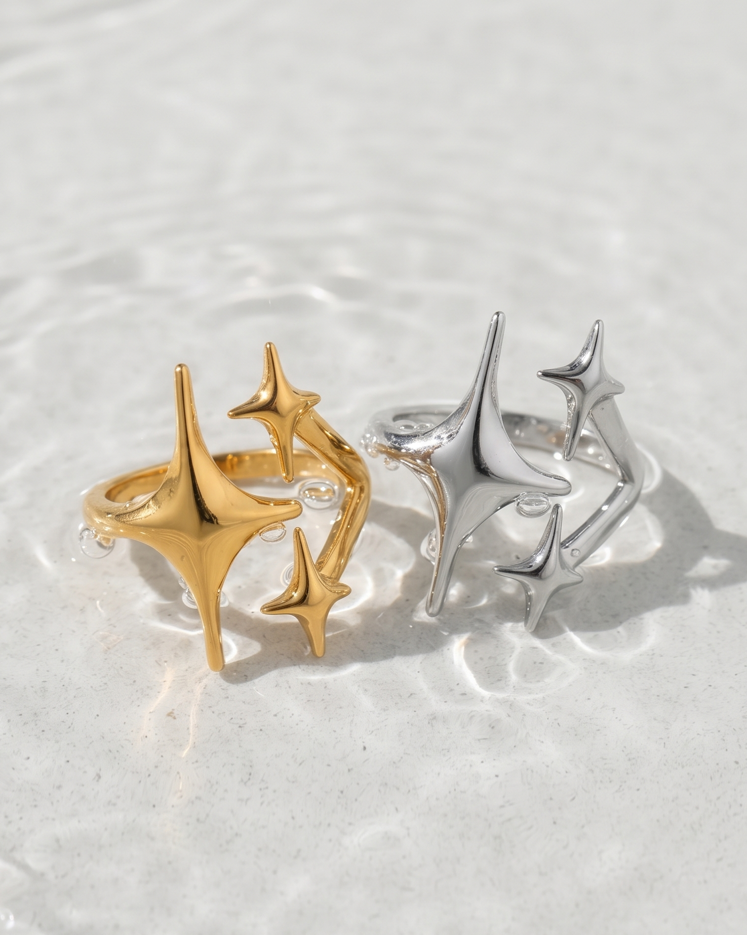 Two star-shaped rings, one in polished gold and one in polished silver, displayed on a textured white surface with soft water ripples.