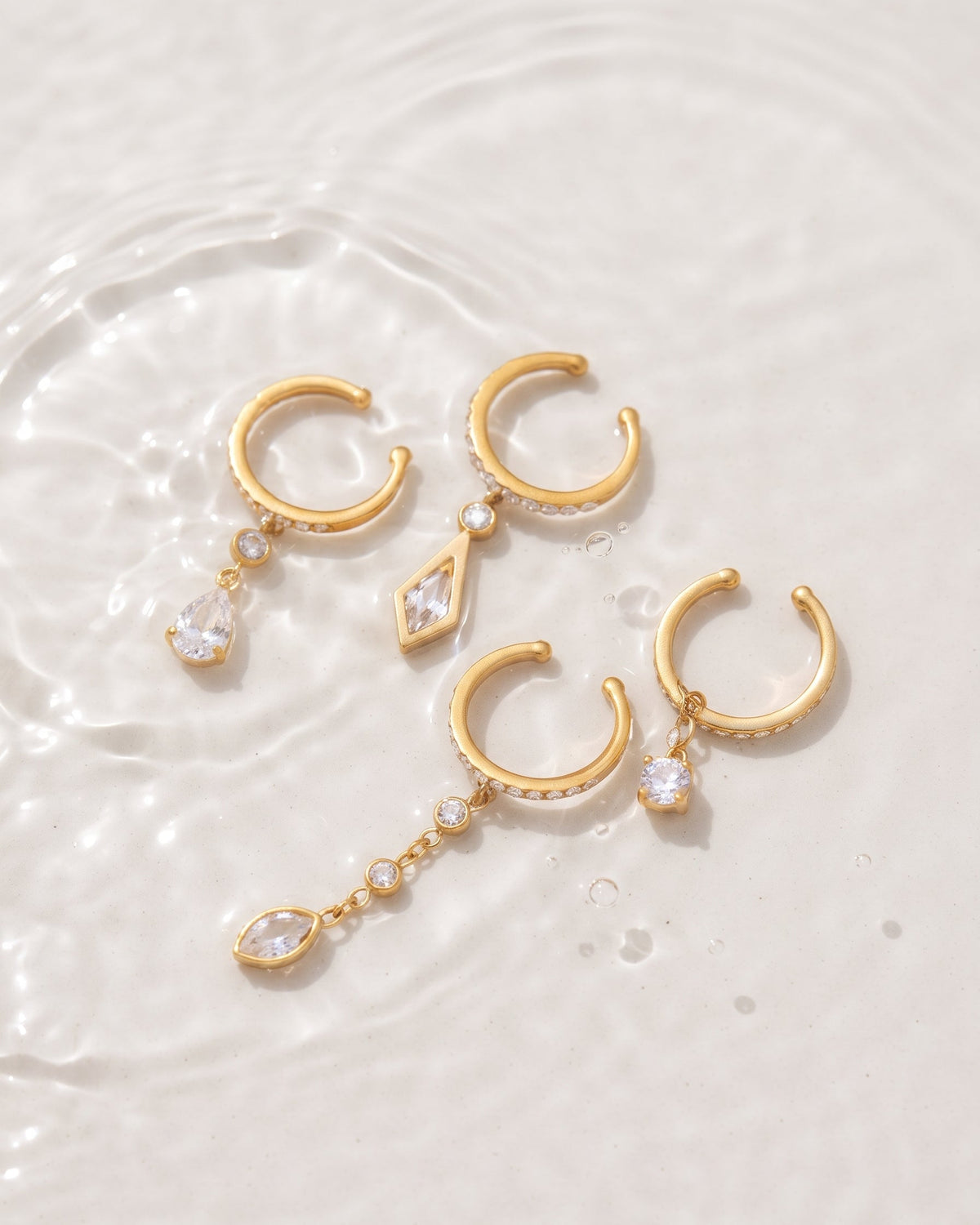 Three gold-toned huggie hoop earrings with dangling clear teardrop and marquise crystals, arranged on a white water surface with ripples.