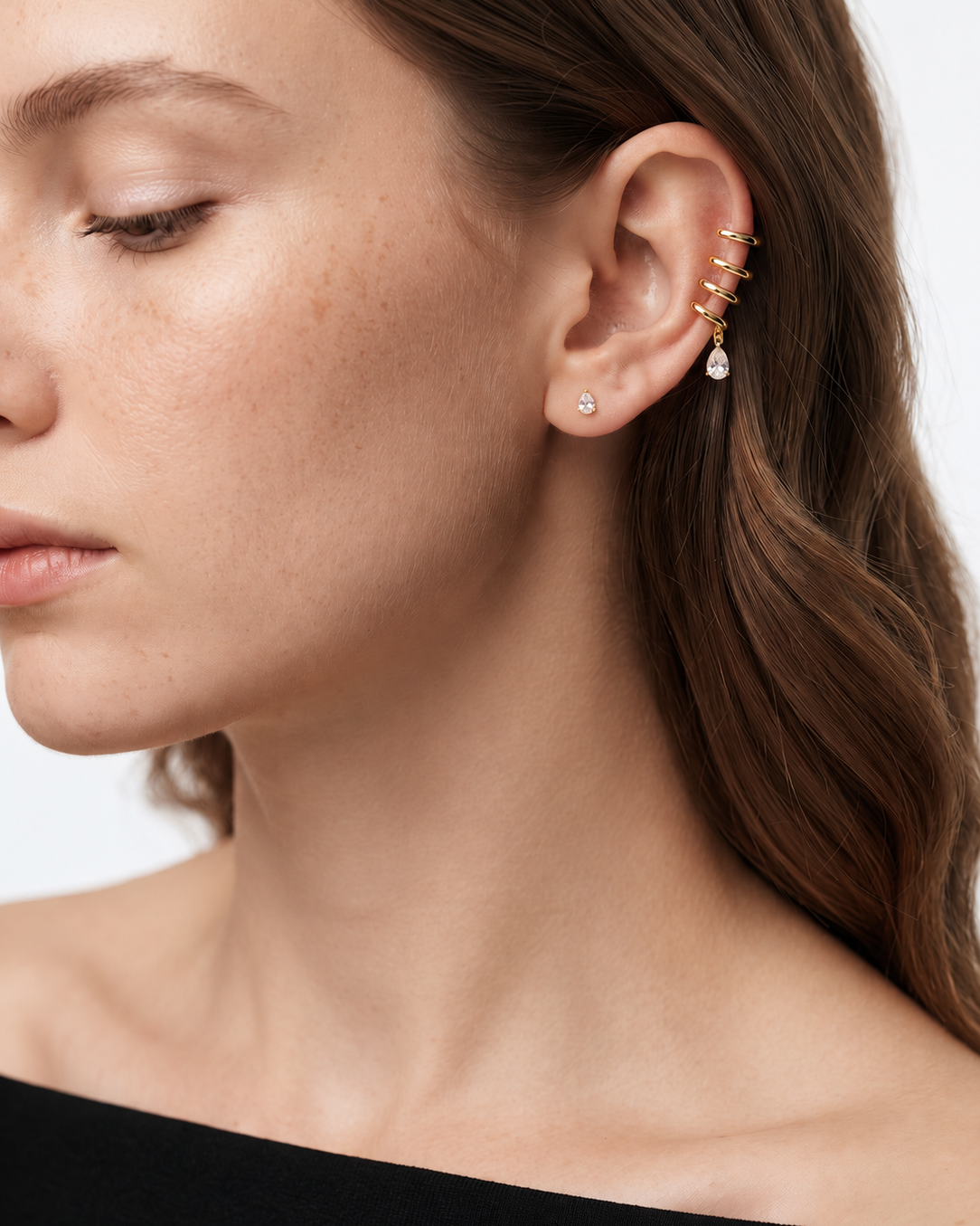 A close-up of a woman's ear wearing a gold ear cuff with small dangling clear stones and a single round stud earring, against a light neutral background.