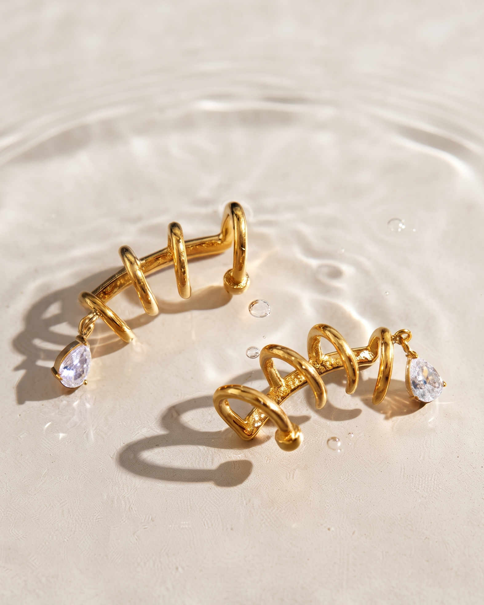 A pair of gold spiral hoop earrings with small clear teardrop gemstones, resting on a light beige surface with water ripples and shadows.