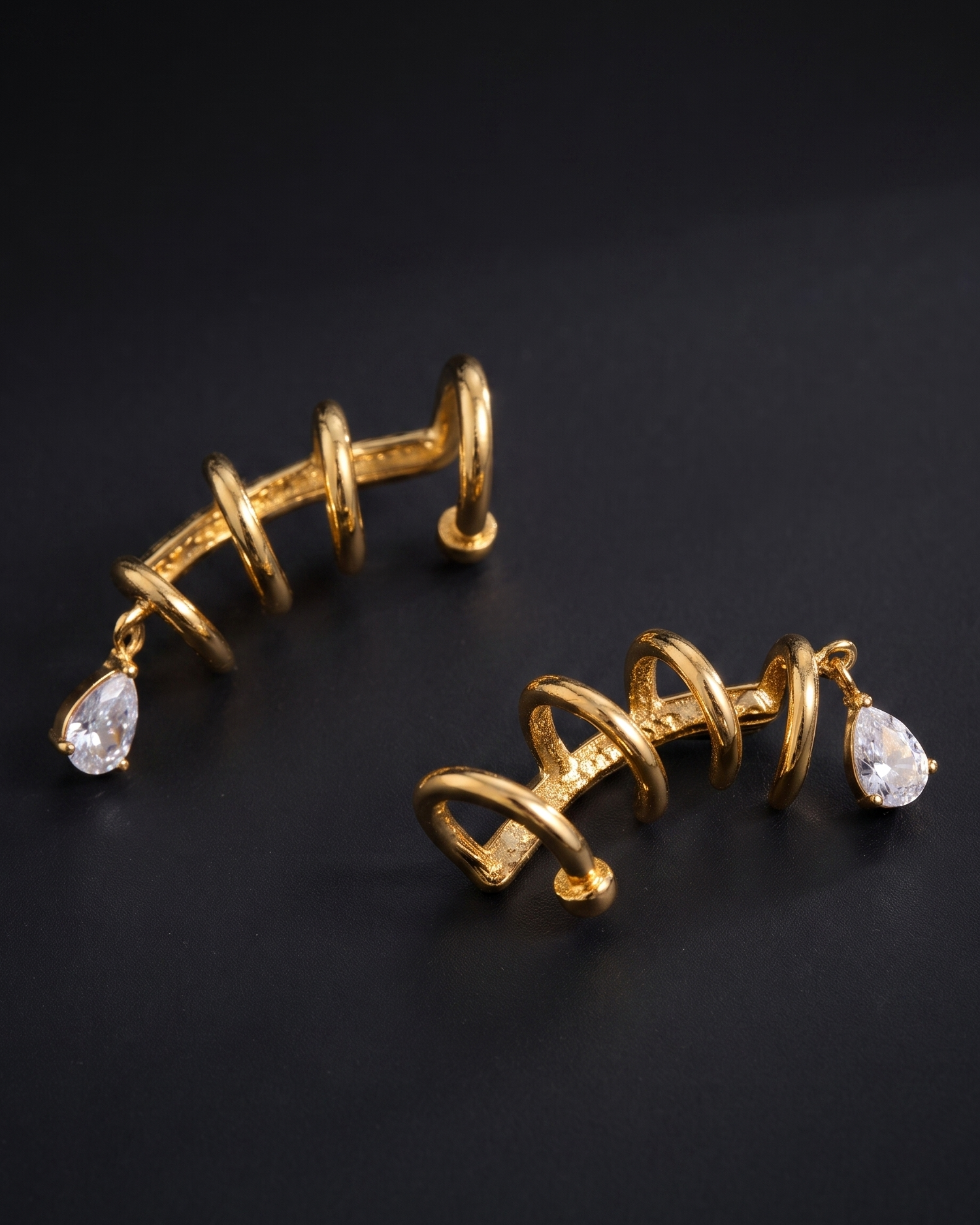 A pair of gold spiral-shaped hoop earrings with teardrop clear gemstones, displayed on a dark black background.