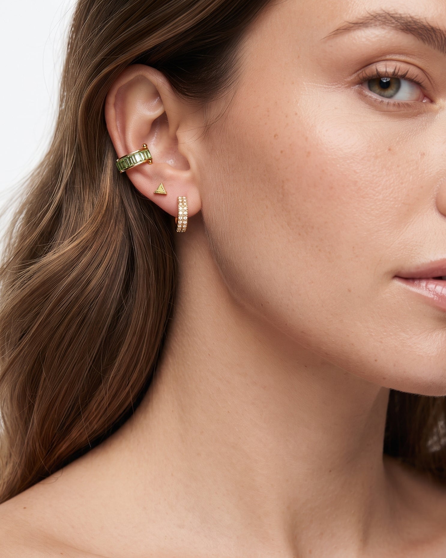 A close-up of a woman's ear wearing multiple gold ear cuffs and studs, with her hair and shoulder visible against a clean light background.
