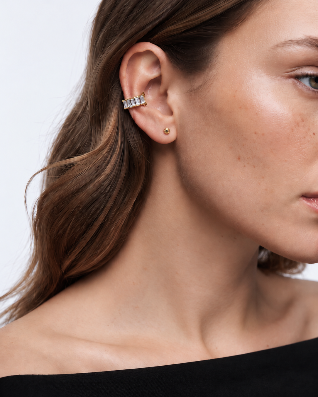 A close-up of a woman's ear wearing a gold ear cuff with clear stones and a small gold stud earring, against a light gray background.