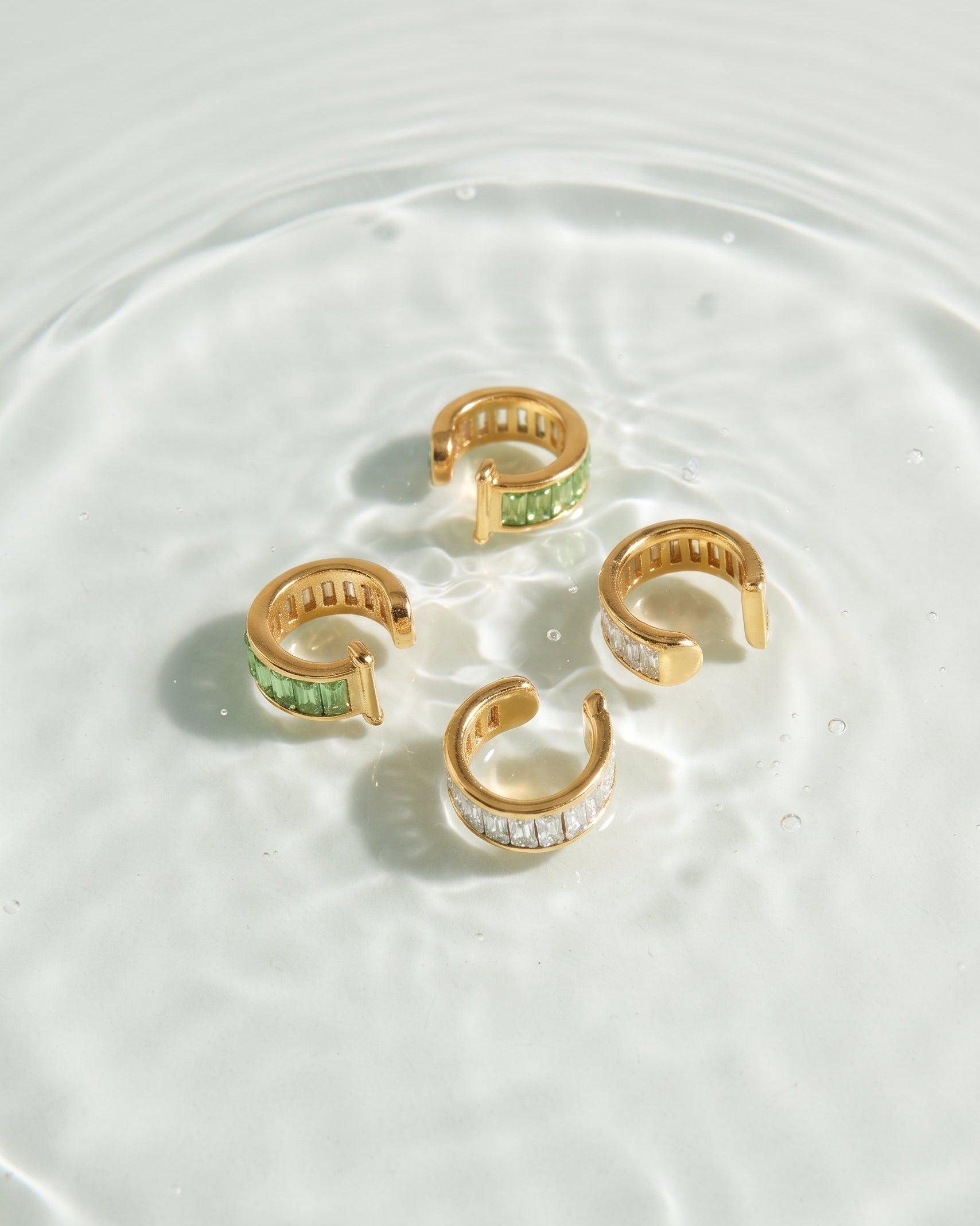 Four gold C-shaped hoop earrings with green and clear gemstone inlays, arranged in a circle on a shallow pool of water.