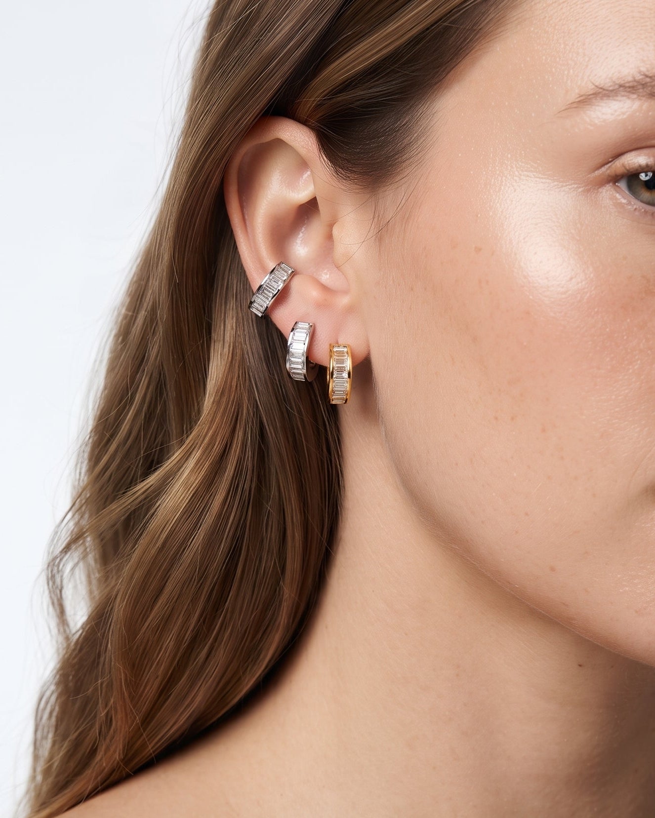A close-up of a woman's ear wearing multiple small hoop and cuff earrings, including silver and gold-toned pieces with clear stones, against a light gray background.