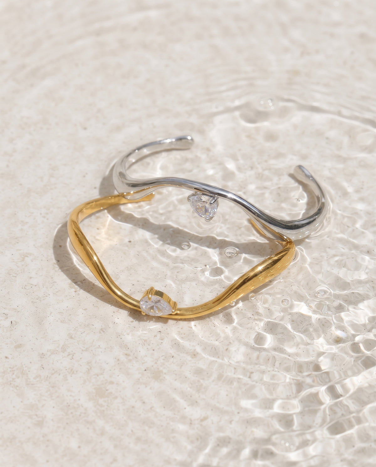 Two open cuff bracelets, one in polished yellow gold and one in polished silver, each featuring a small clear heart-shaped gemstone, resting on a textured light beige surface with water ripples in the background.