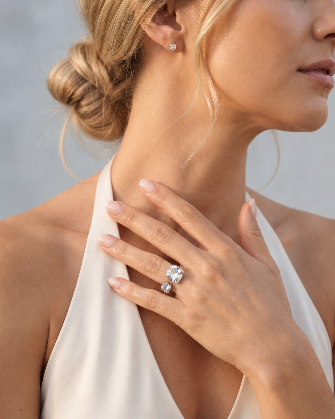 A close-up lifestyle shot of a woman wearing a silver ring with a large clear oval gemstone and a matching small stud earring. The ring is shown on her finger, with her hand resting near her collarbone against a soft gray background.