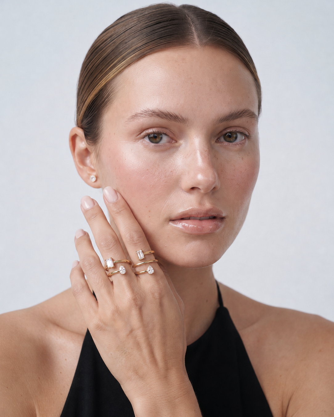 A close-up portrait of a woman with her hand resting against her cheek, showcasing a stack of delicate gold rings with small clear gemstones. She is wearing a black halter top against a clean, light gray background.