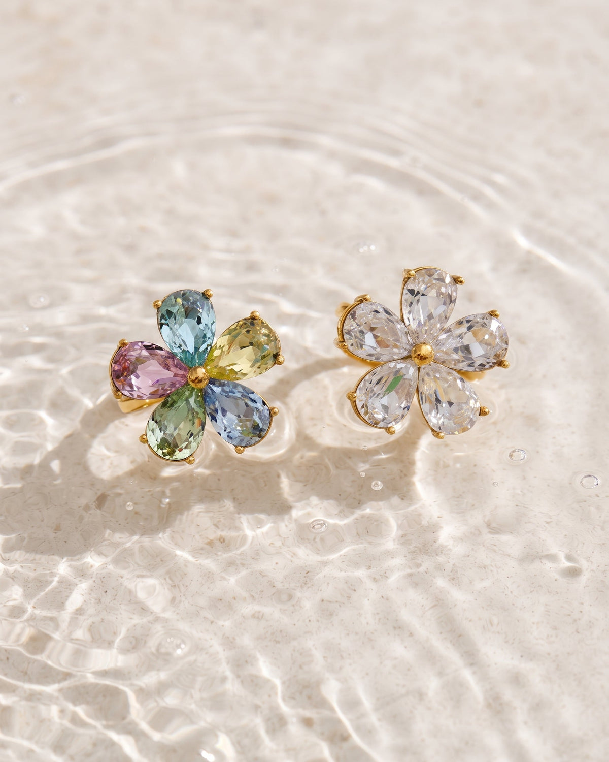 A pair of gold flower-shaped stud earrings resting on shallow water. One earring features multicolored pastel gemstones (blue, green, pink, yellow), while the other features clear, colorless gemstones.
