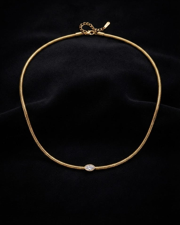 A delicate gold snake chain necklace with a small oval clear gemstone at the center, displayed on a black velvet background.
