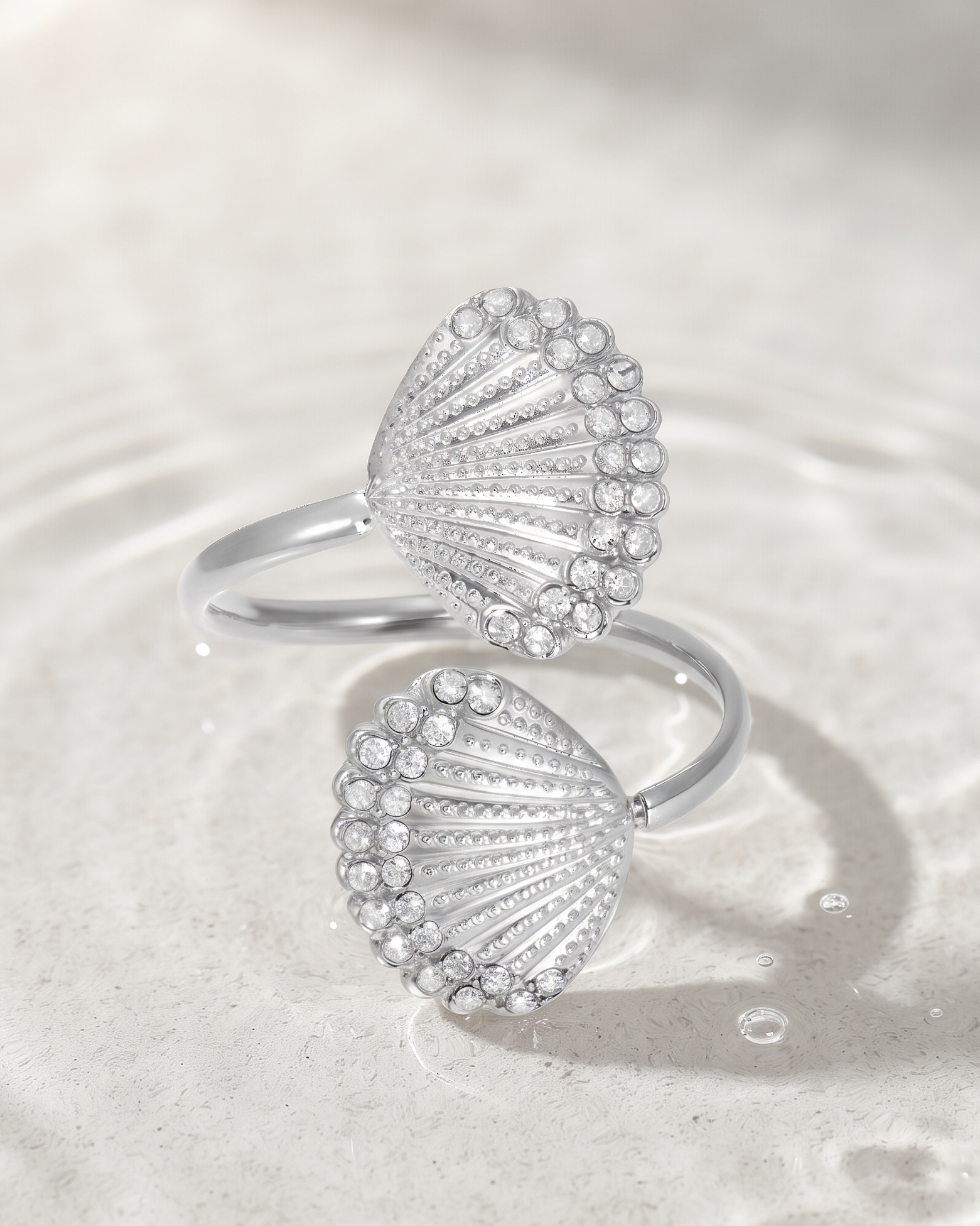 A pair of silver-toned shell-shaped rings set with clear stones, resting on a light beige textured surface with subtle water ripples.