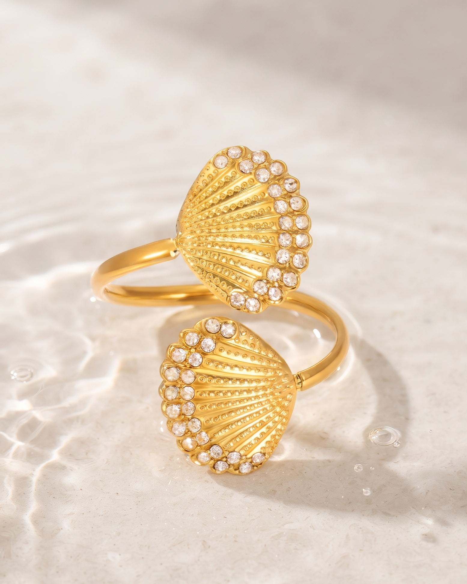 Two gold seashell-shaped rings with small clear stones along the shell edges, displayed on a light beige textured surface with soft water ripples and shadows.