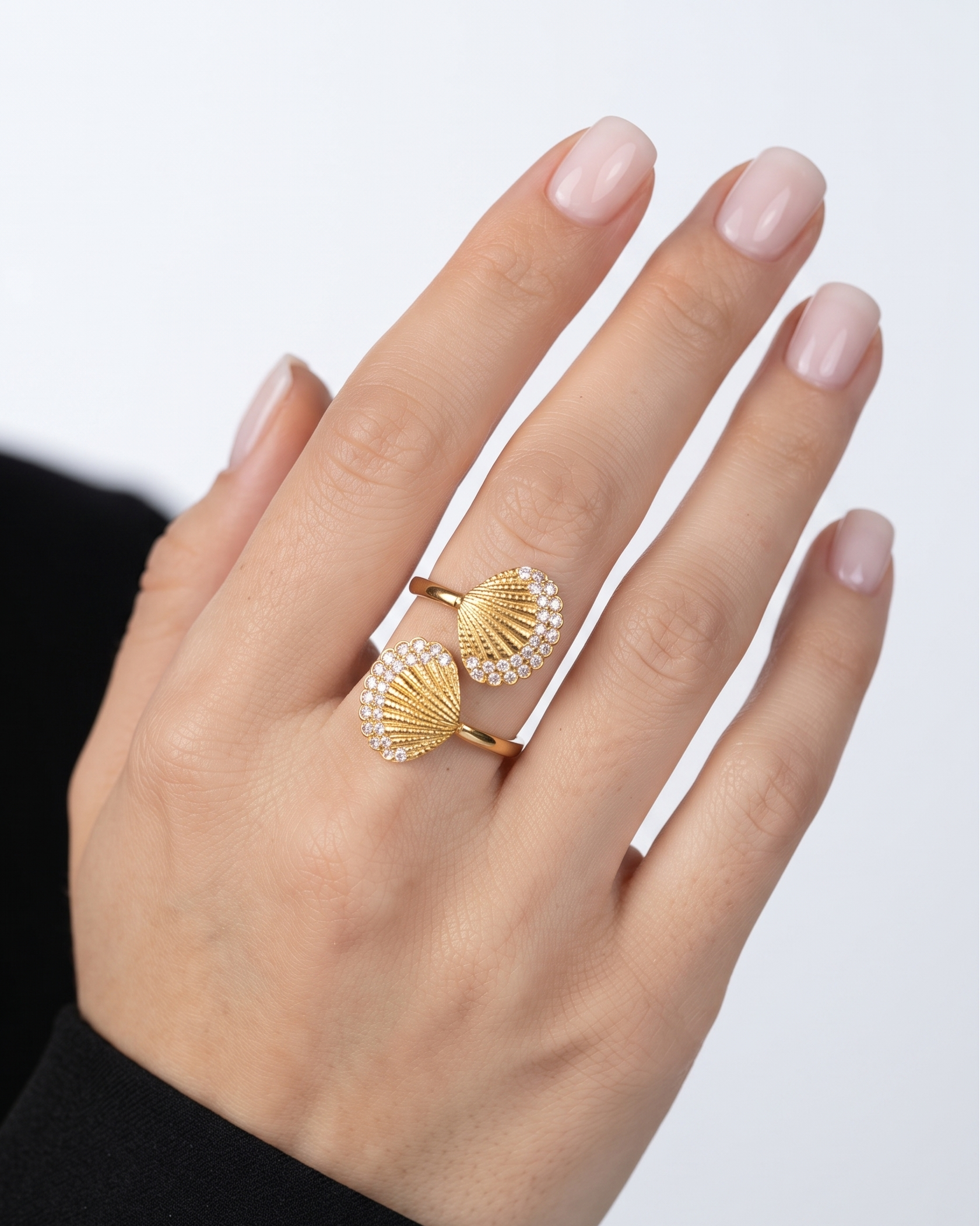 A close-up of a hand wearing two gold rings with fan-shaped designs set with small clear stones, against a light gray background.