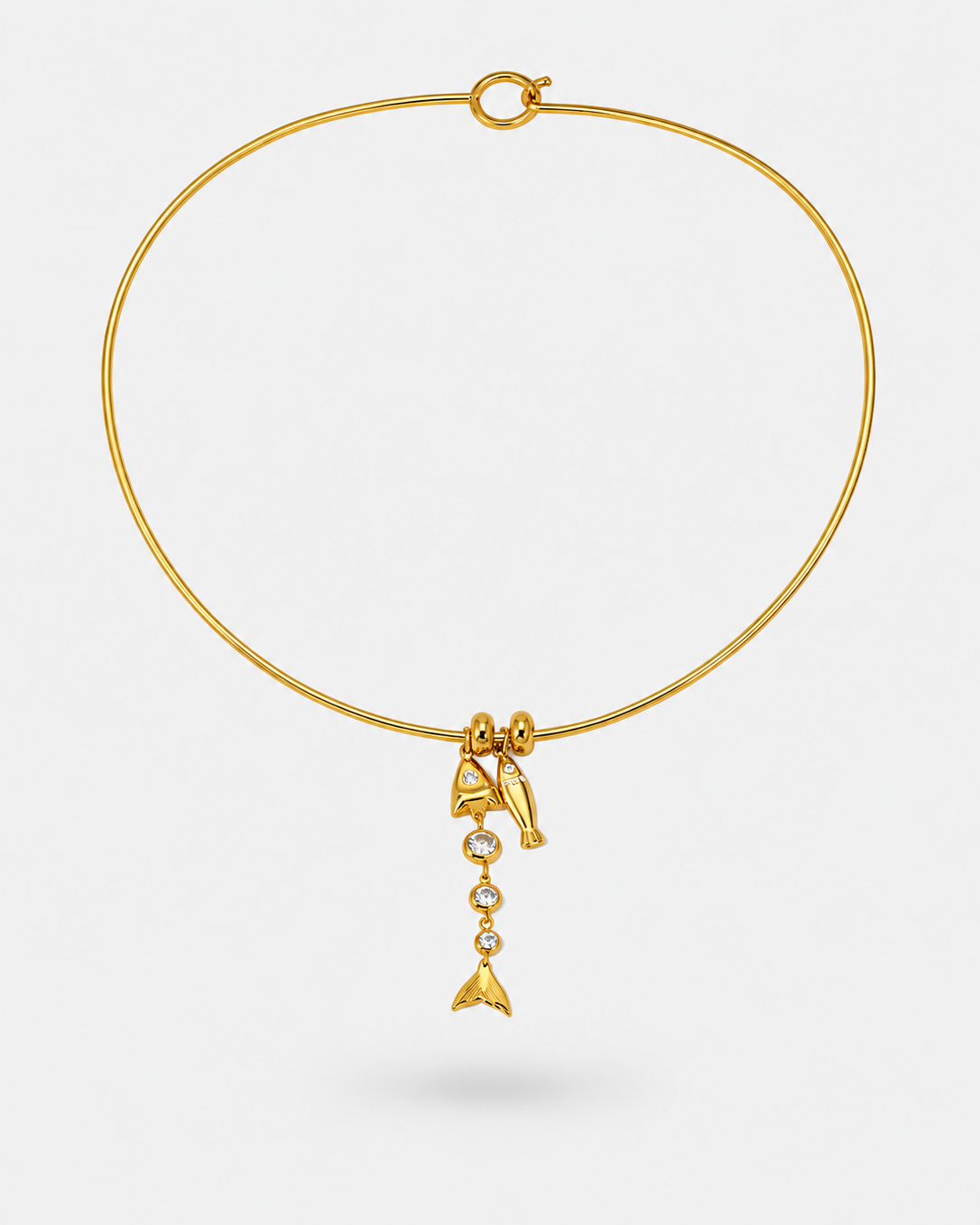 A delicate gold wire bangle bracelet with a small dangling charm featuring a bow, a small gold fish, and a tiny dangling pearl-like bead, shown on a light gray background.