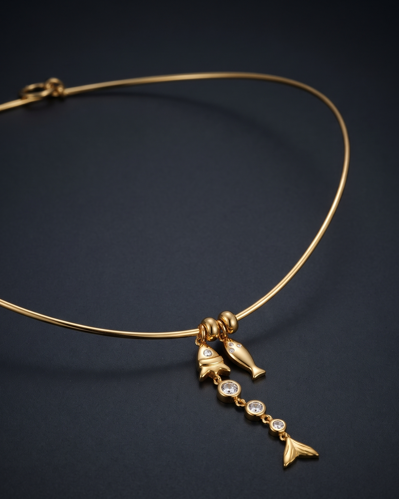 A delicate gold wire necklace with a dangling charm featuring two dolphins, a small shell, and two clear gemstones, displayed on a dark textured background.