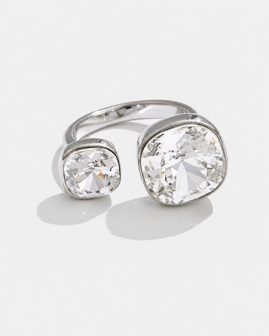 A silver-toned open ring featuring two cushion-cut clear gemstones in bezel settings, displayed on a light gray background.