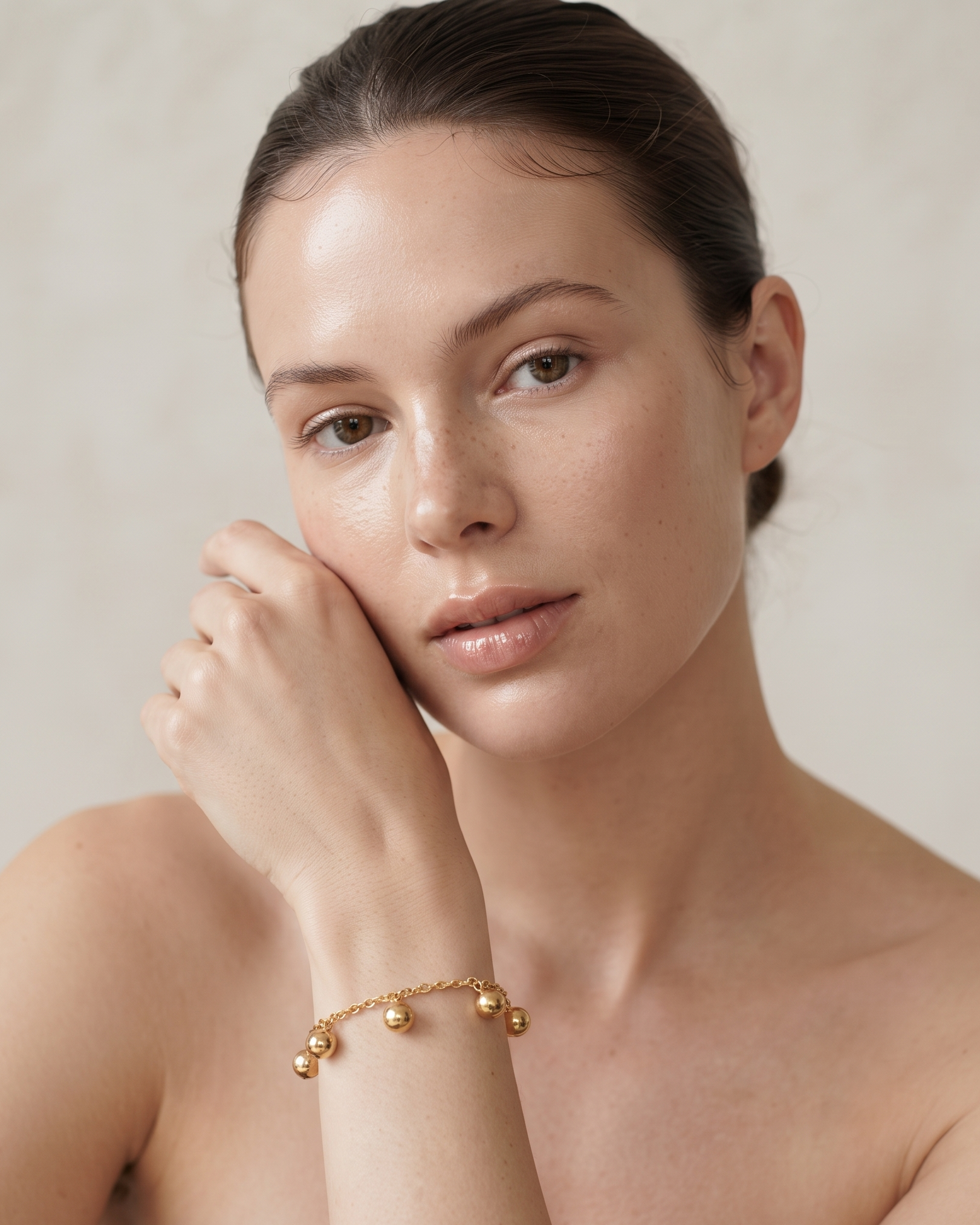 A close-up portrait of a woman wearing a delicate gold chain bracelet with small bead charms, posed against a soft beige background.