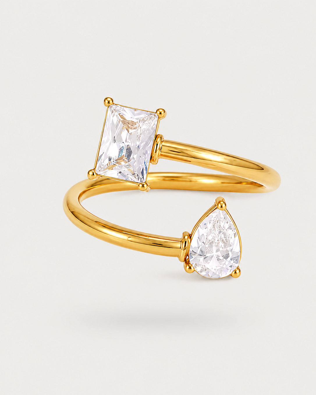 A gold ring featuring a rectangular clear gemstone and a pear-shaped clear gemstone on a slim gold band, set against a light gray background.