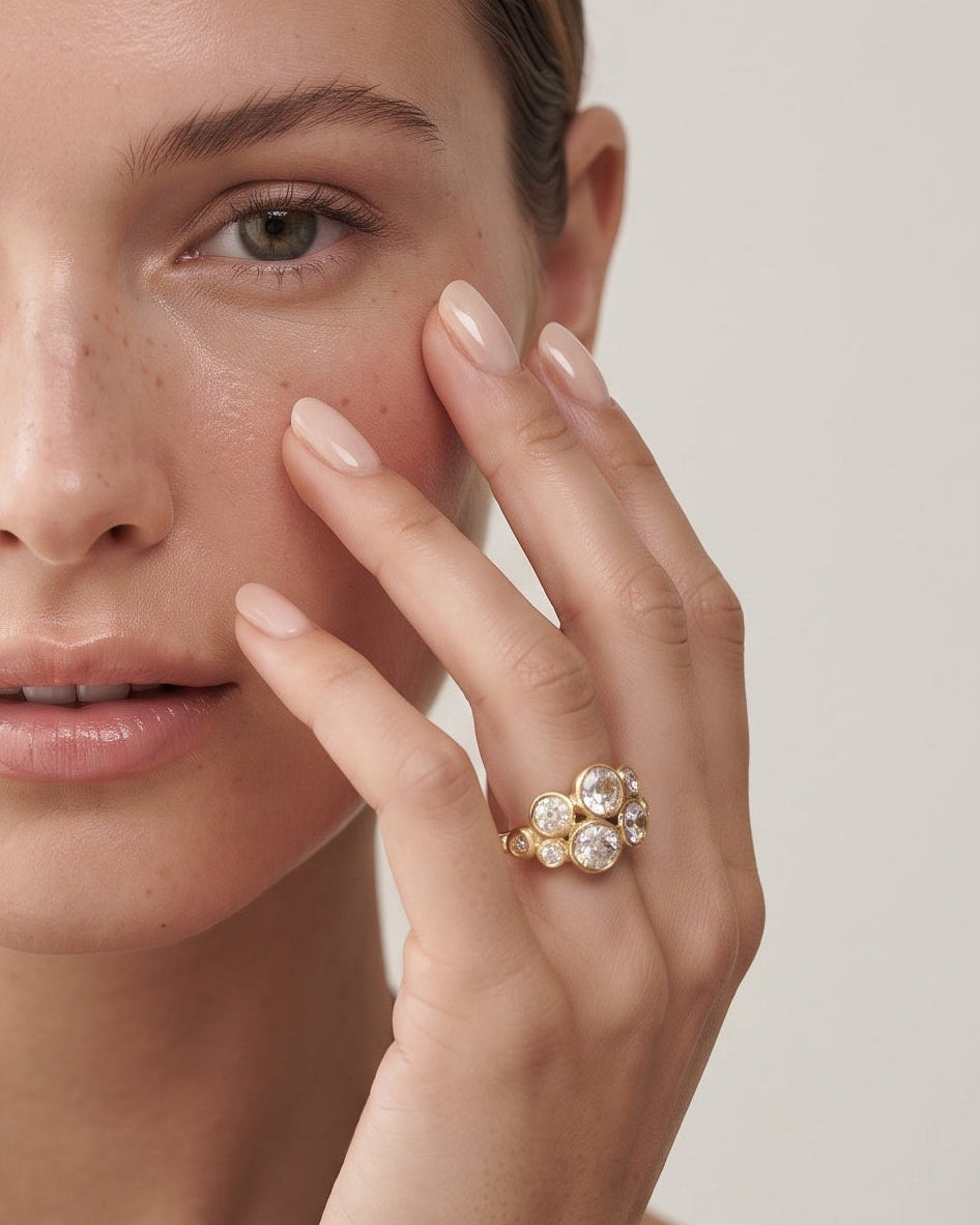 A close-up portrait of a woman wearing a gold ring with clear gemstones on her finger, set against a soft, light neutral background.