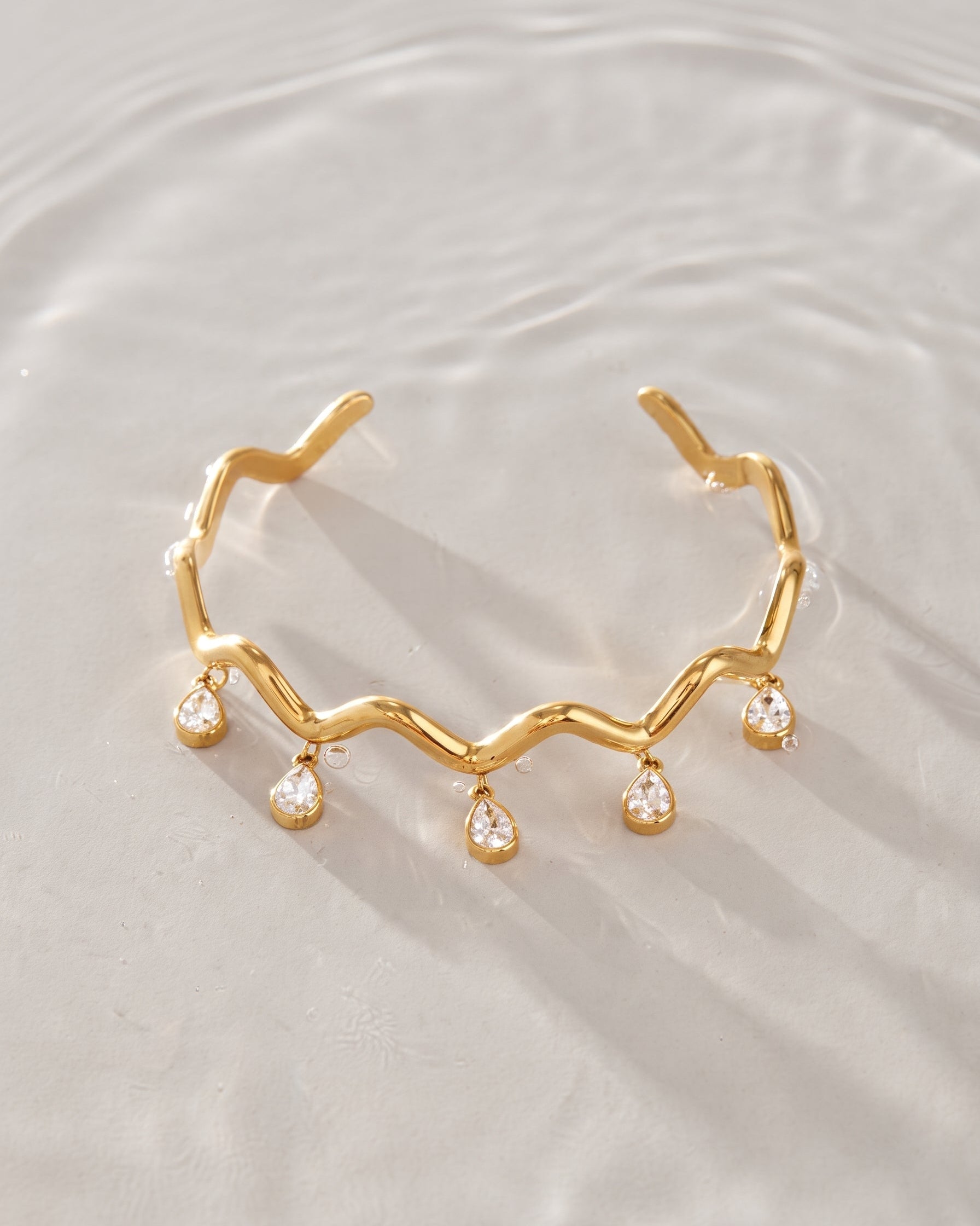 A gold open cuff bracelet with a wavy zigzag line and five dangling teardrop-shaped clear gemstones, photographed on a softly rippled light gray surface.