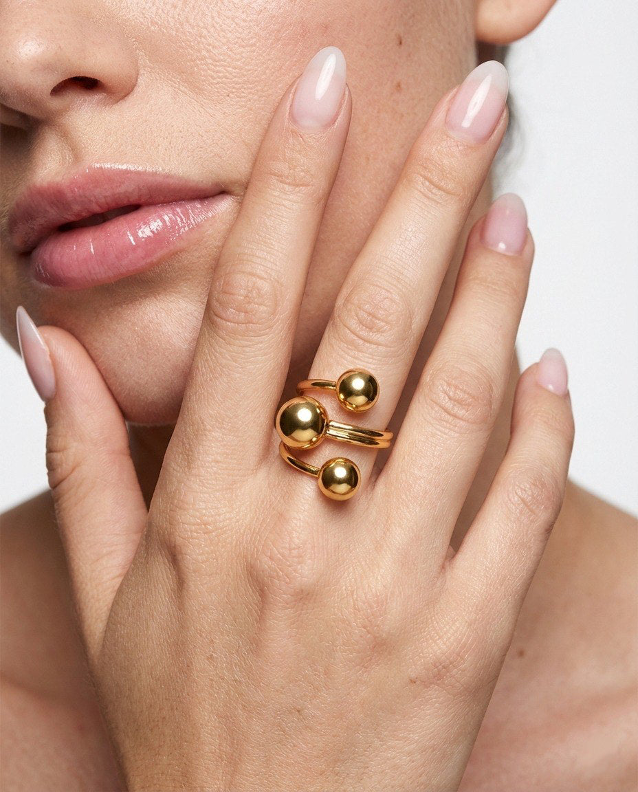 Close-up of a hand wearing a gold ring with three spherical elements, against a neutral background.