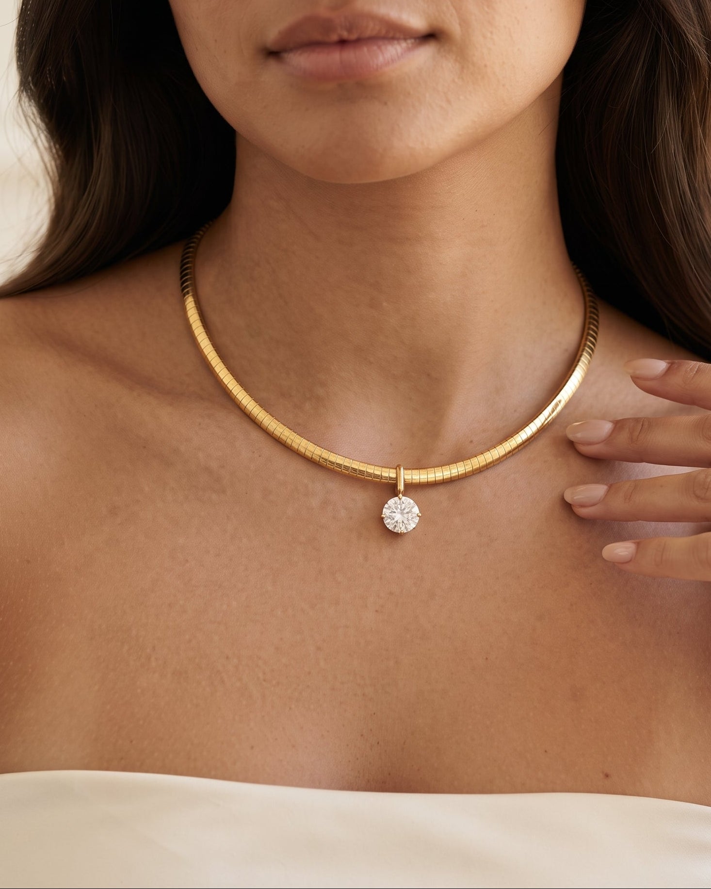 A close-up of a woman wearing a textured gold snake chain necklace with a small round clear gemstone pendant, paired with a white strapless top.