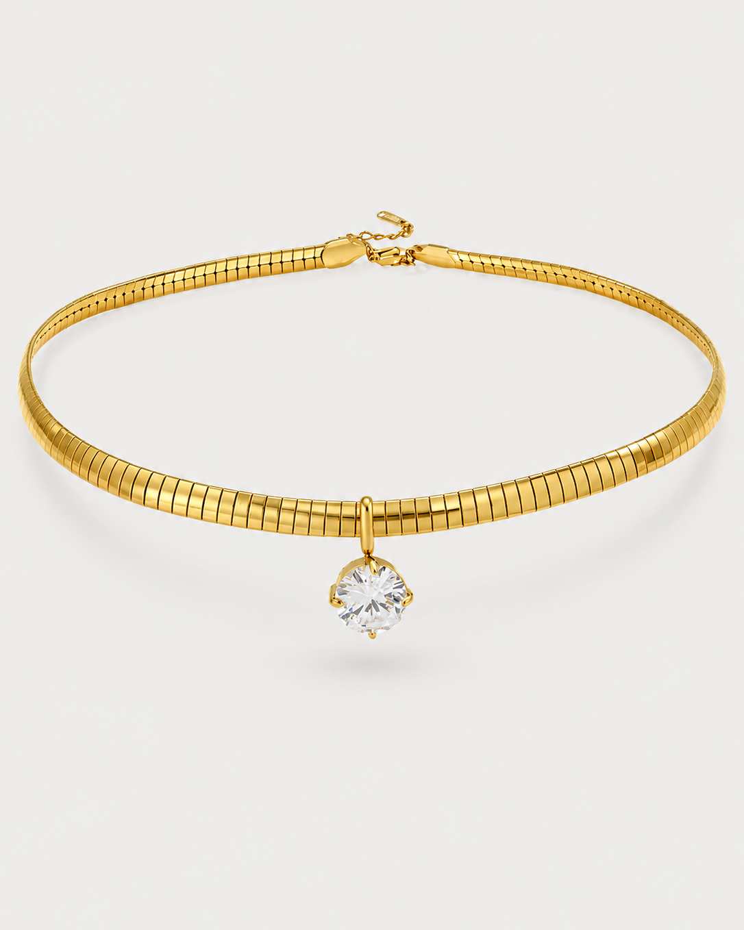 A gold snake chain choker necklace featuring a single bezel-set clear round gemstone pendant, displayed on a light gray background.